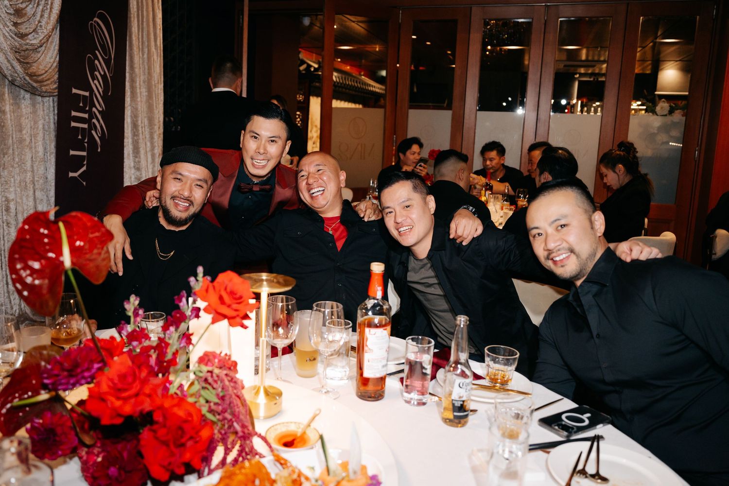 Five men in black attire smile at a festive dinner table with red floral arrangements and whiskey bottles.