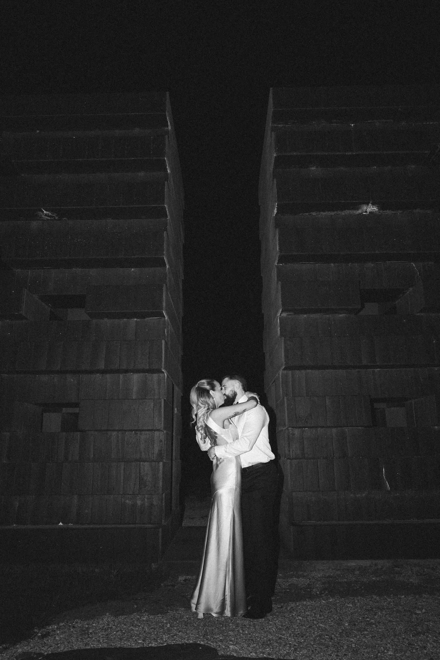 A dramatic black and white wedding photo of a couple embracing between tall industrial structures.