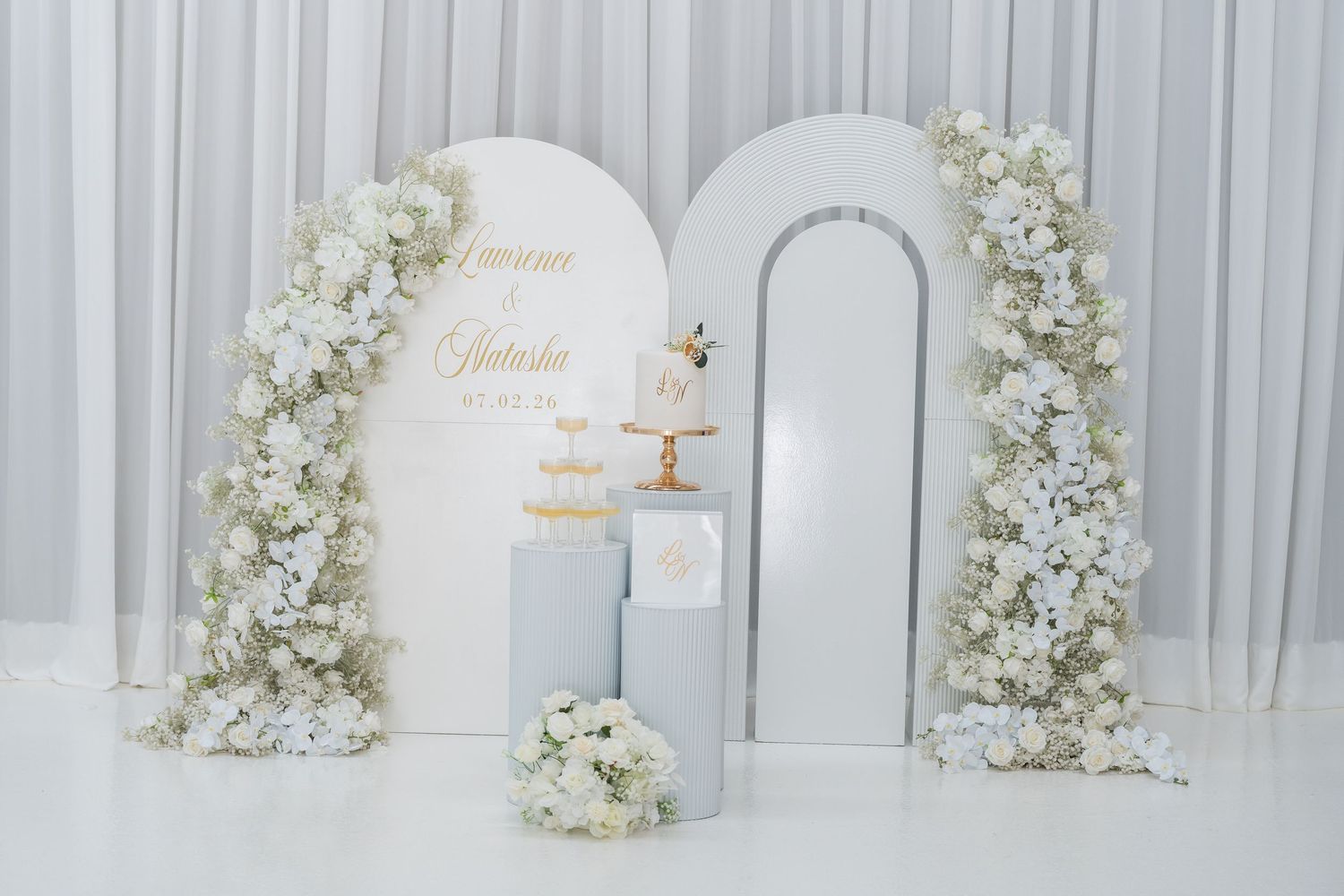 Elegant wedding backdrop with white floral arch, cake display, and personalized name sign in soft gold lettering.