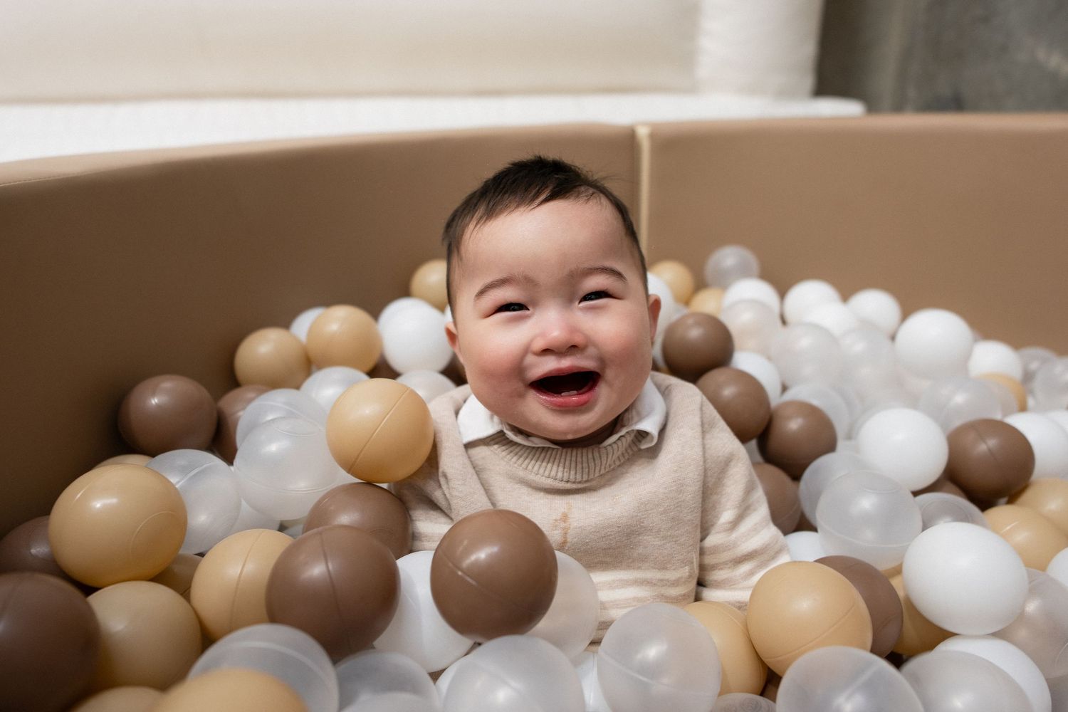 Happy baby laughing in a cardboard box ball pit filled with brown, gold, and white plastic balls.
