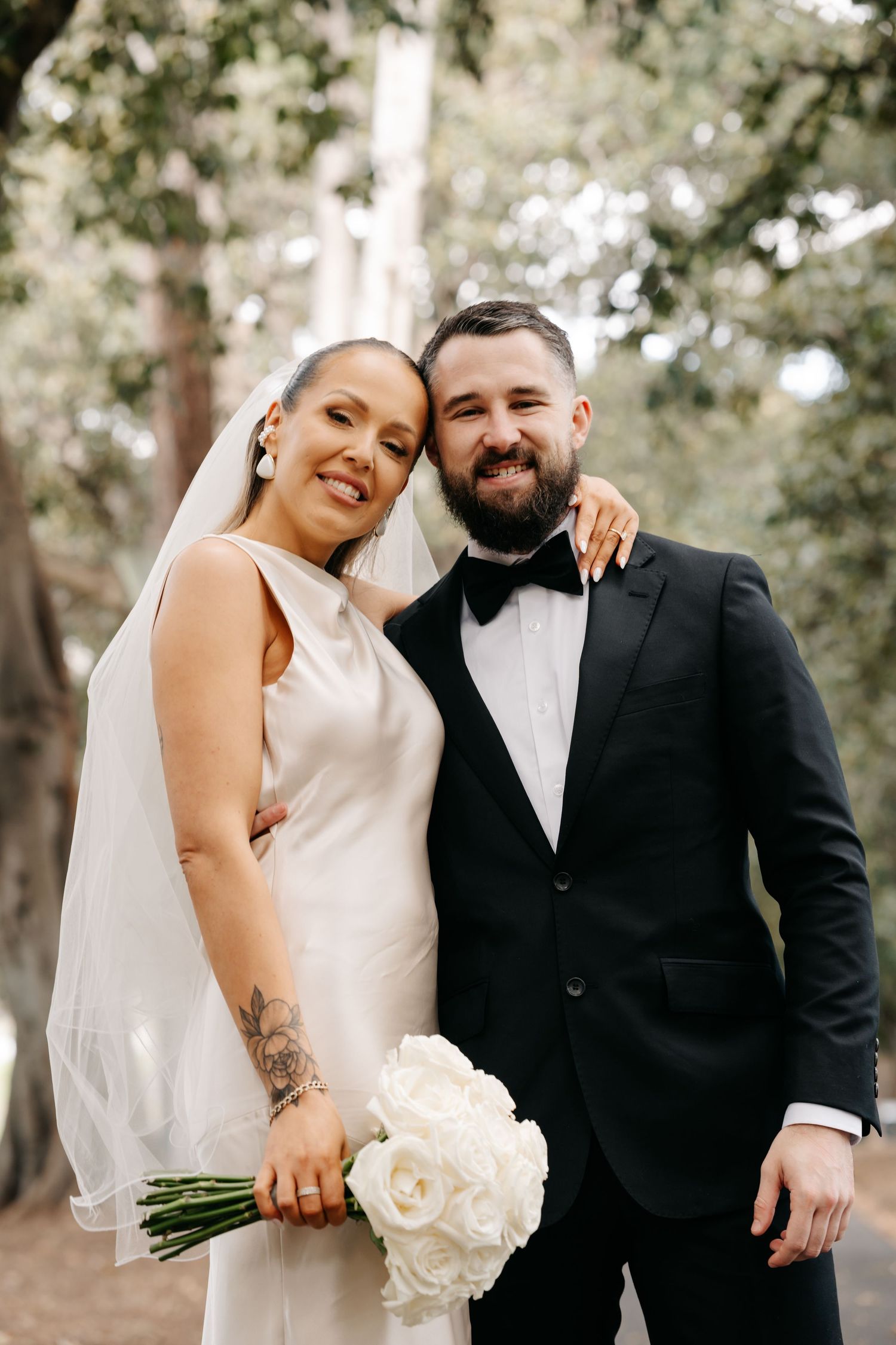 Wedding couple poses together in a wooded outdoor setting with white floral bouquet.