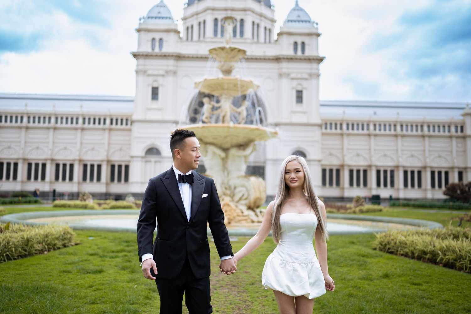 A couple strolls hand in hand in formal attire in front of a grand historic building with fountain.