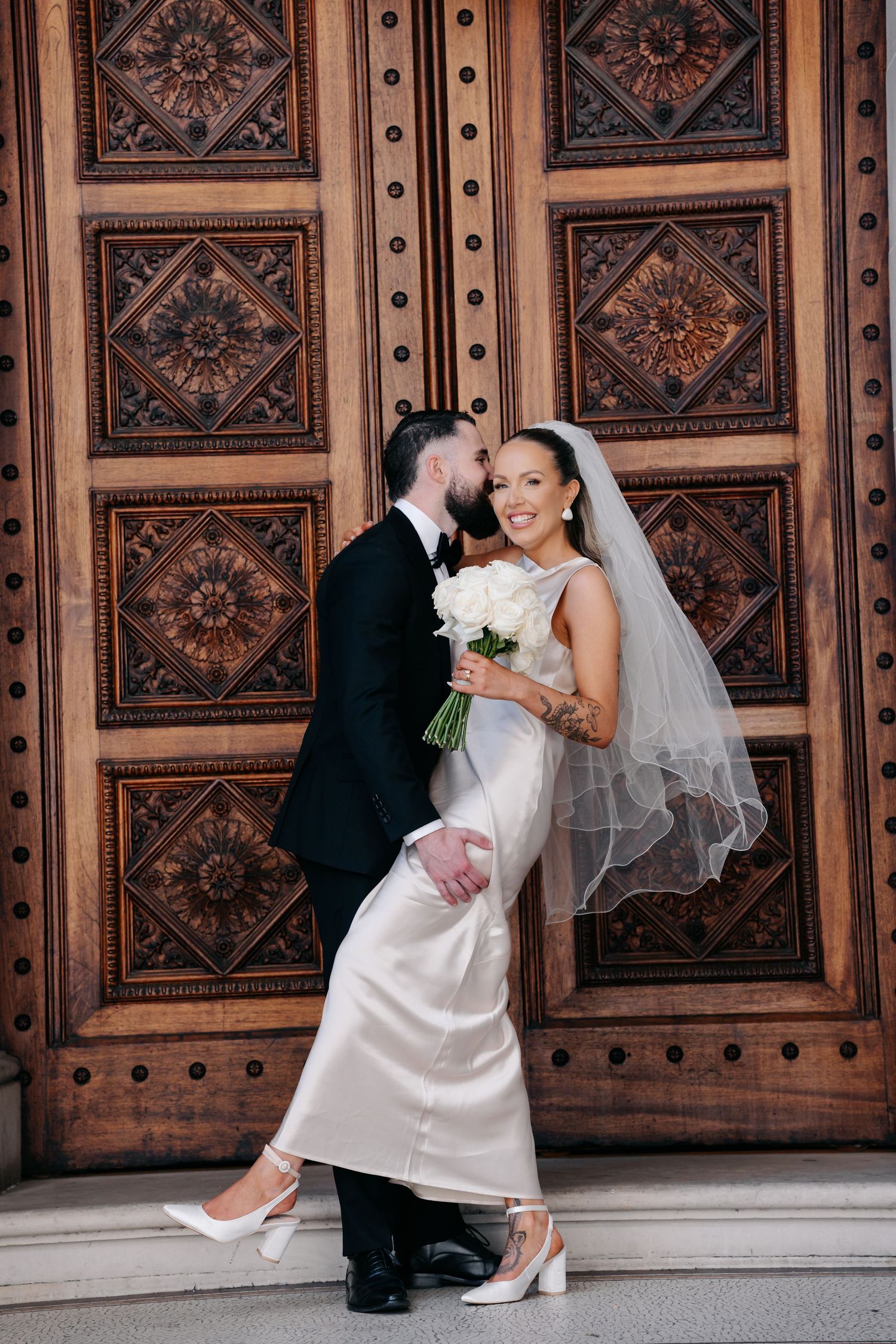 Stylish wedding portrait against ornate wooden door showing bride's elegant white dress.