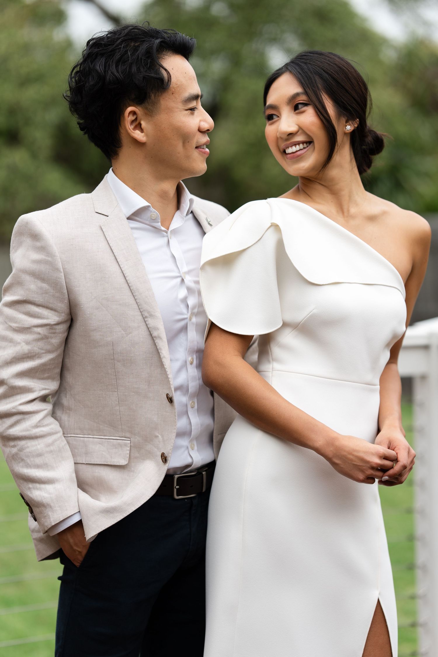 Asian couple in wedding attire, groom in beige suit and bride in white one-shoulder dress, smiling outdoors.