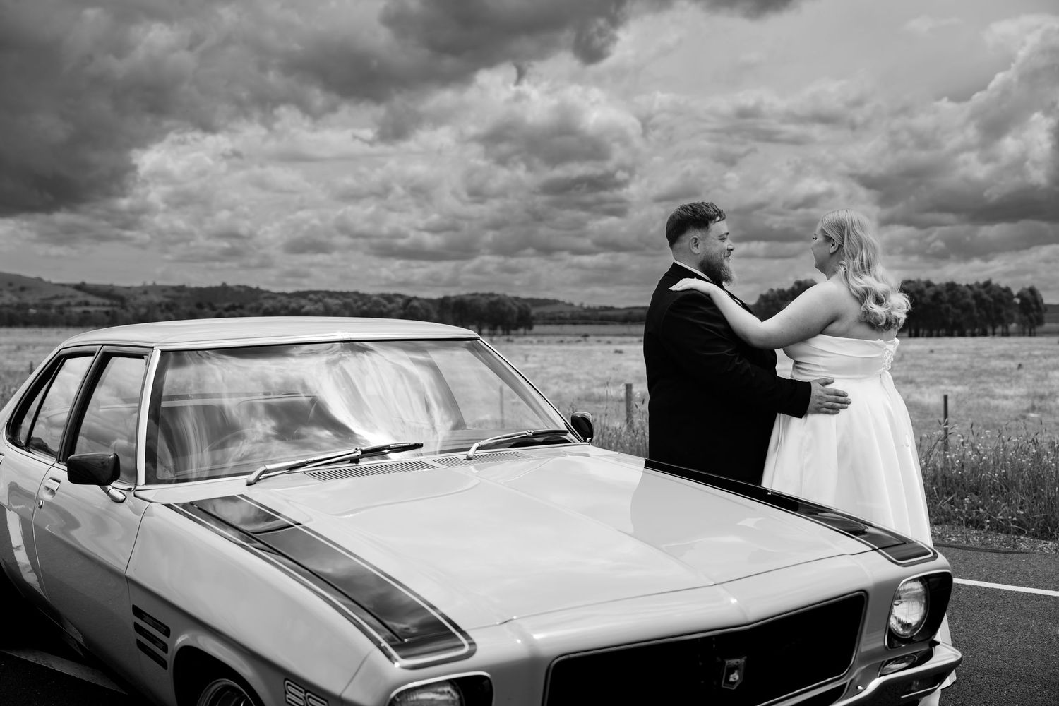 Black and white photo of a bride and groom embracing beside a classic muscle car under dramatic cloudy skies.