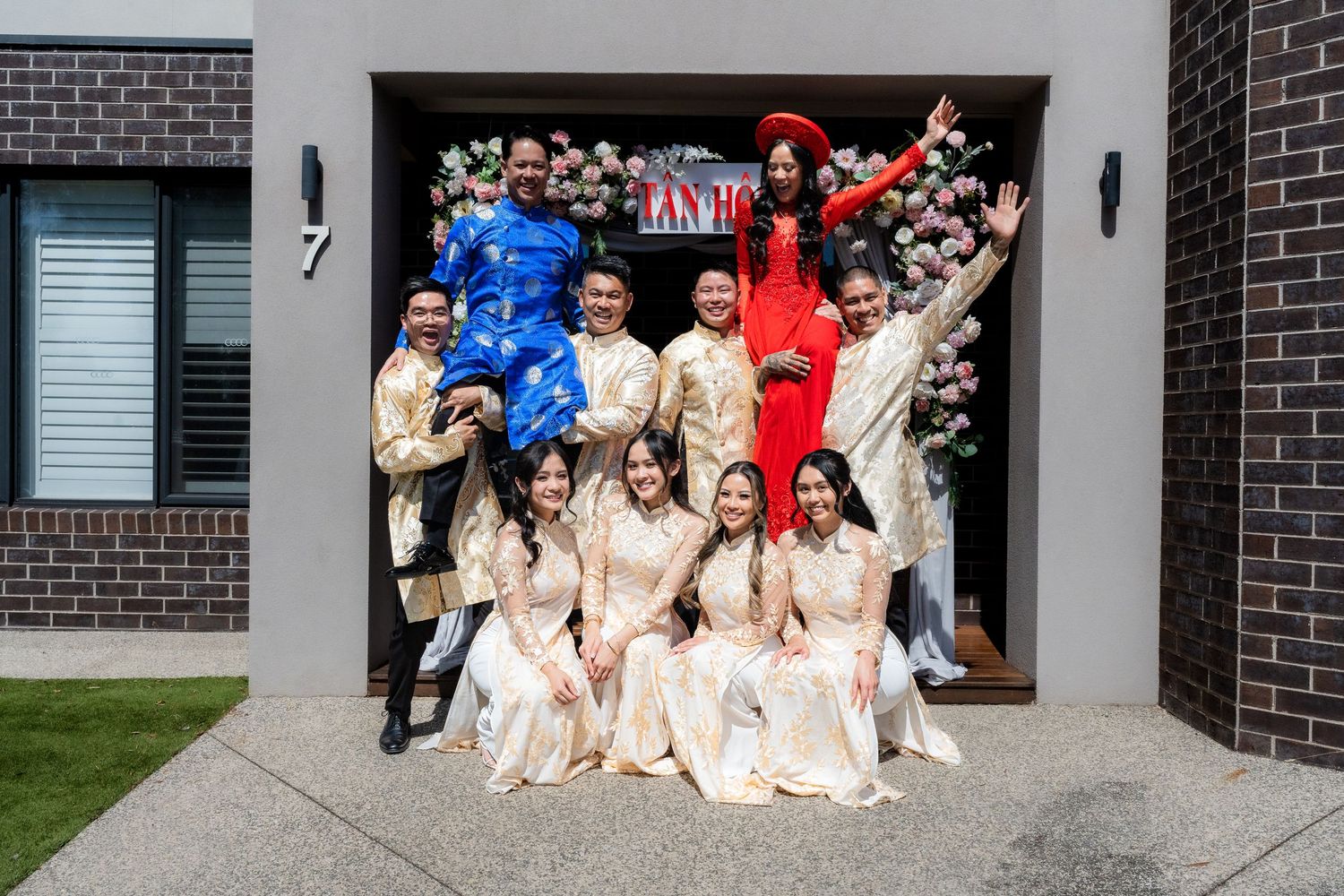 Vietnamese wedding party in traditional ao dai attire posing joyfully outside a modern brick home.
