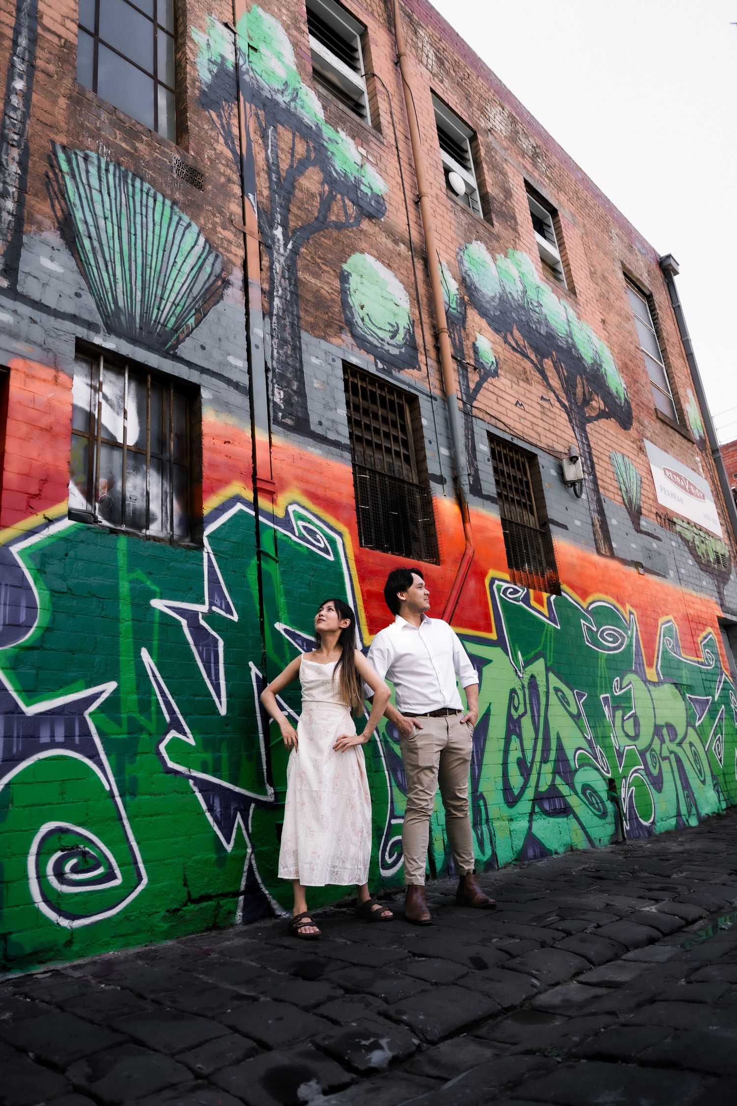 Couple standing before colorful graffiti mural on brick building in urban alley, woman in white dress, man in casual attire.