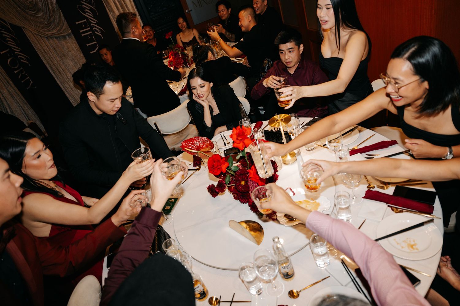 Elegant dinner party guests raising glasses in a toast at a round table with red floral centerpiece.