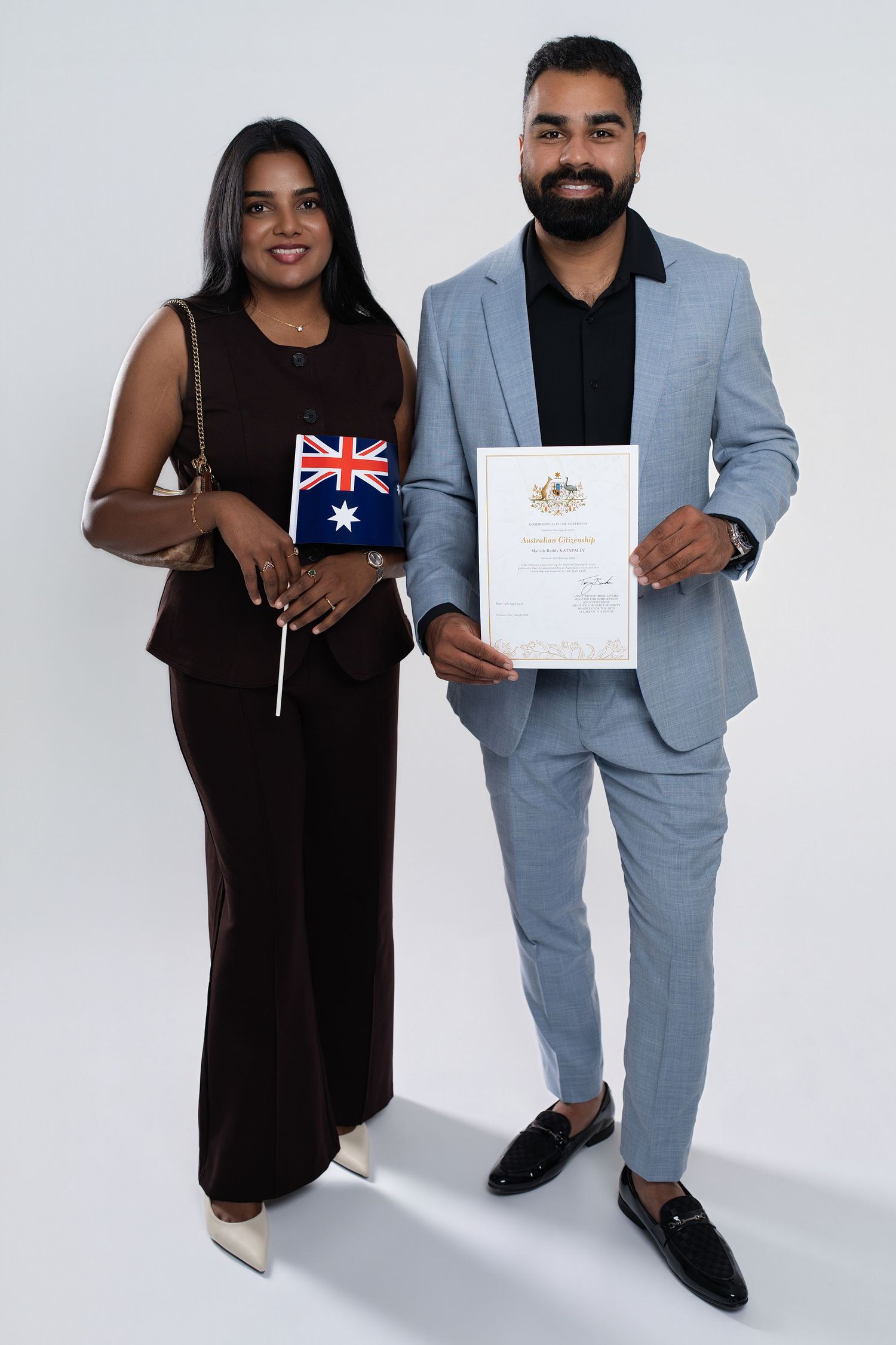 Couple proudly holding Australian flag and citizenship certificate in formal attire against white background.