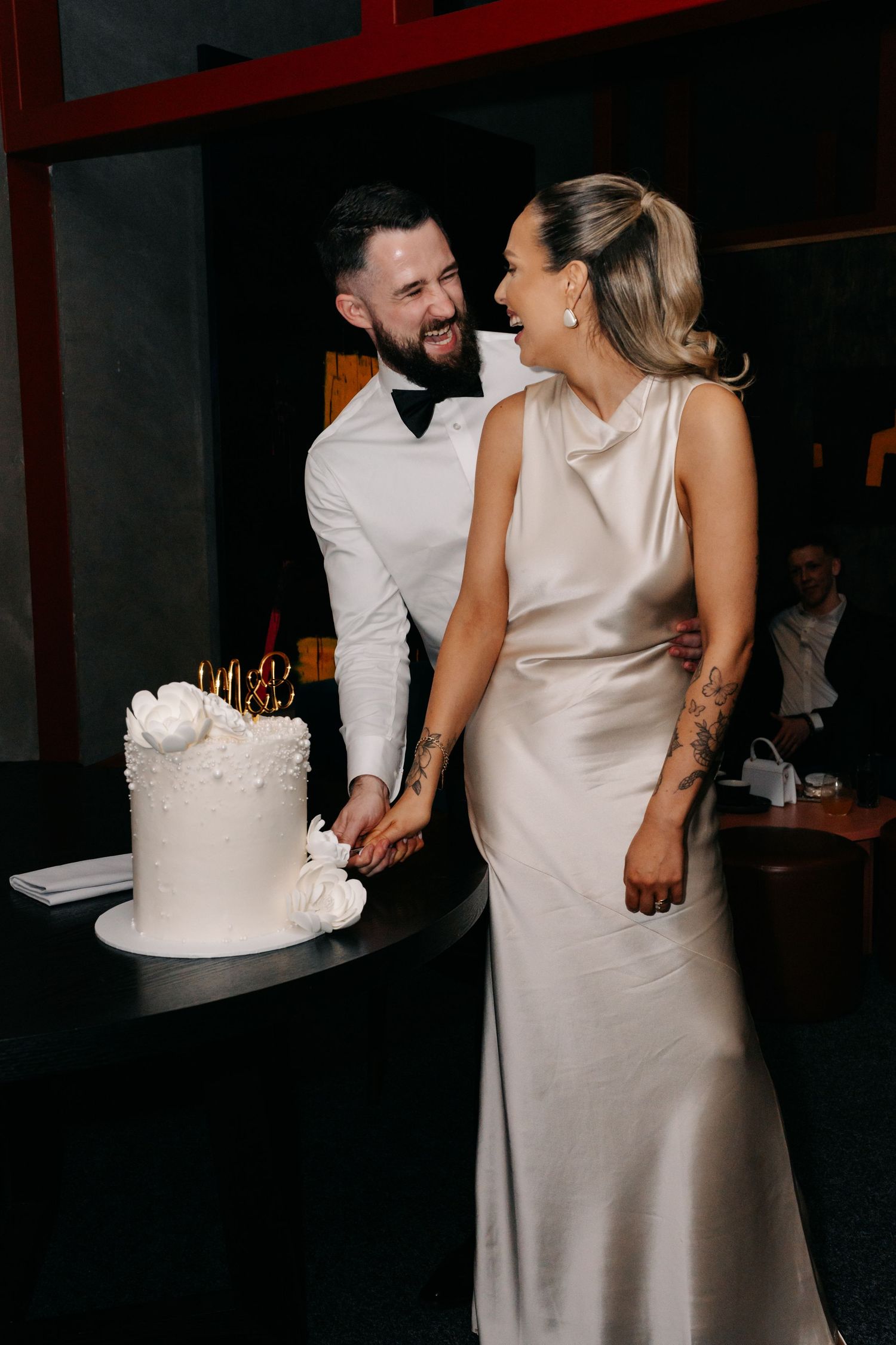 A joyful couple sharing a laugh while cutting their white wedding cake together.