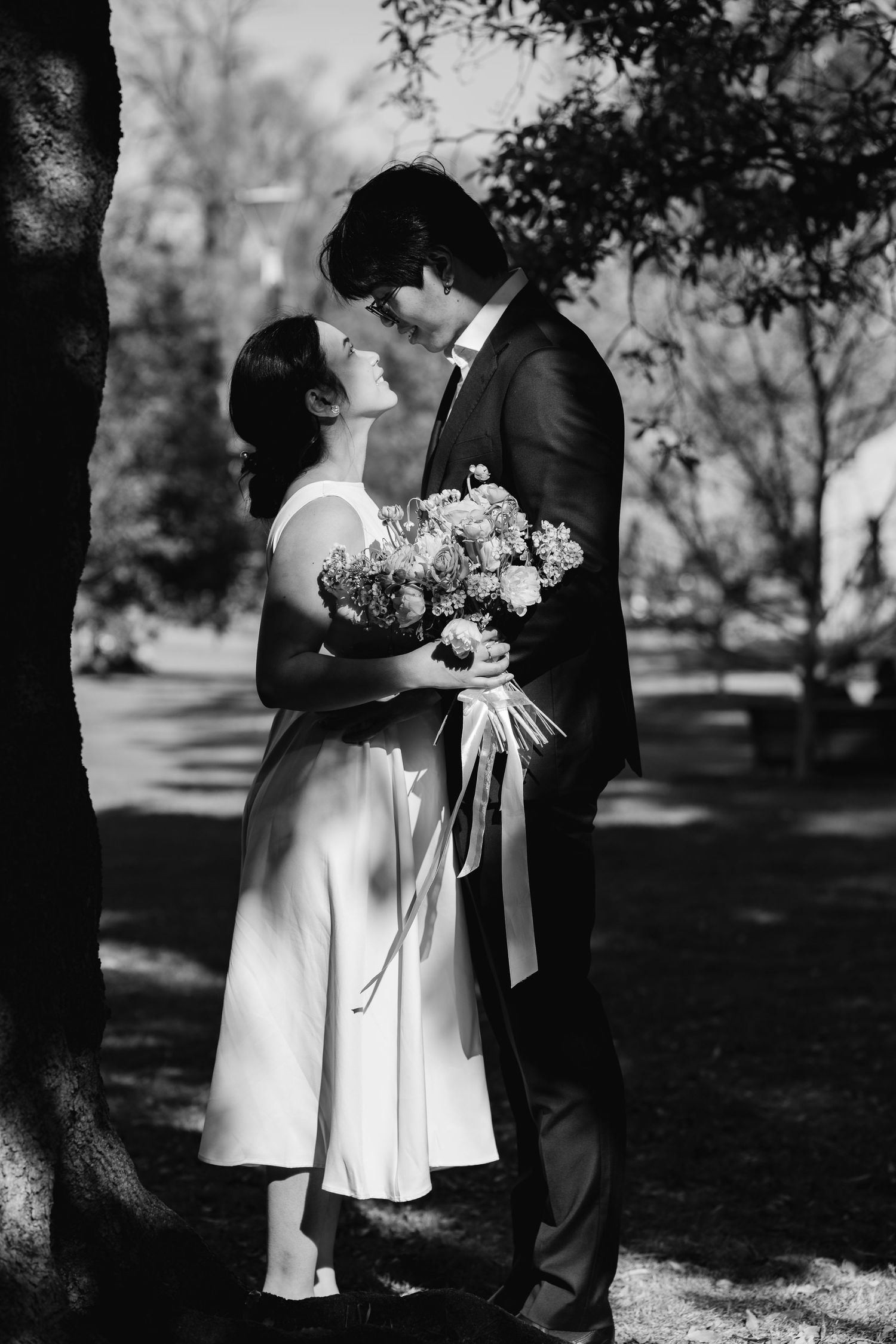Black and white portrait of couple with floral bouquet sharing intimate moment under tree.