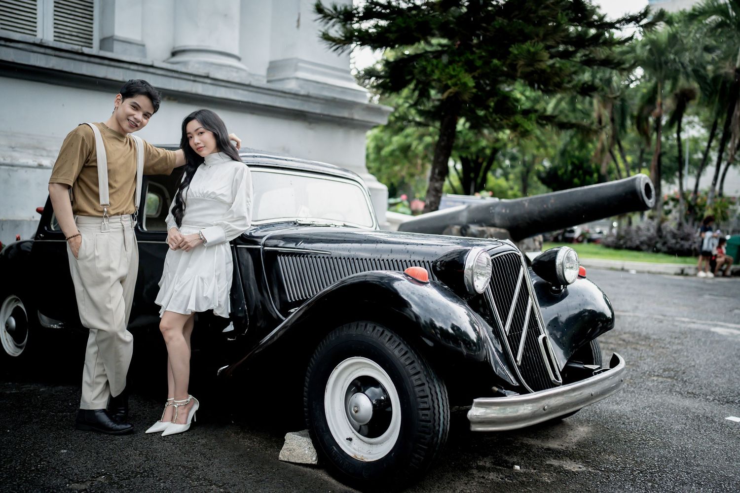 A vintage black car is featured in an elegant photoshoot with a couple near a white colonial building.