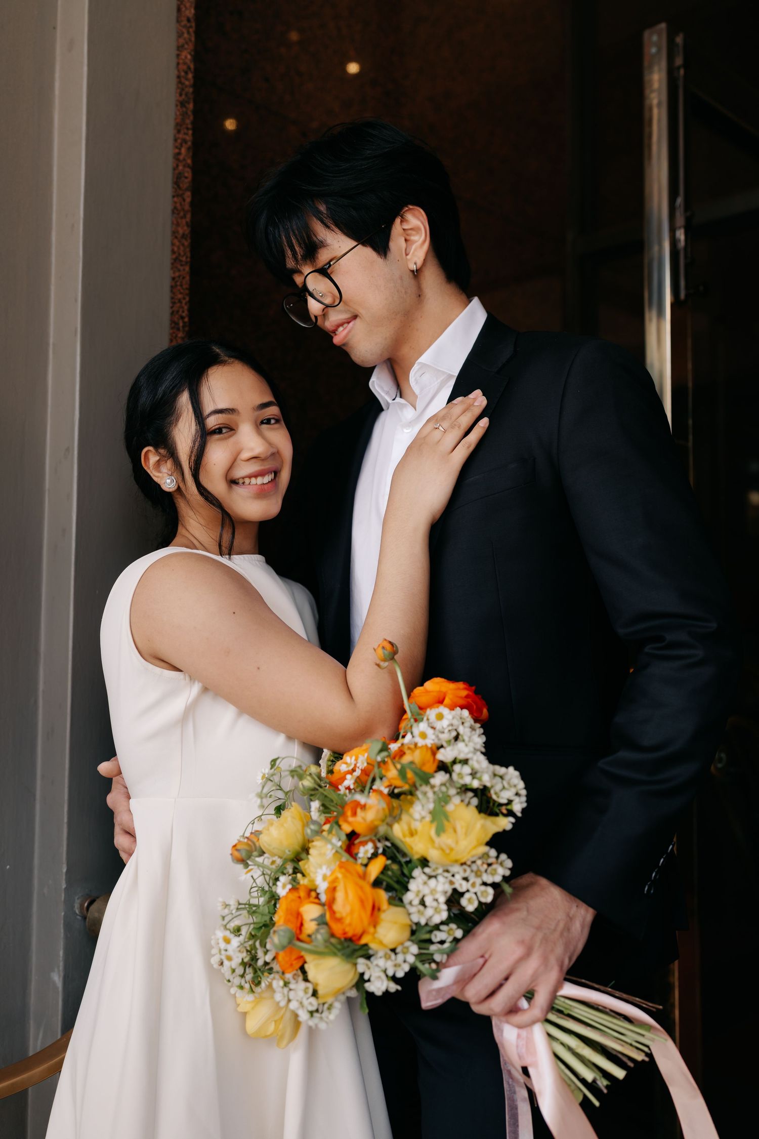 Couple shares tender moments while holding yellow and orange flower bouquets near hotel entrance.