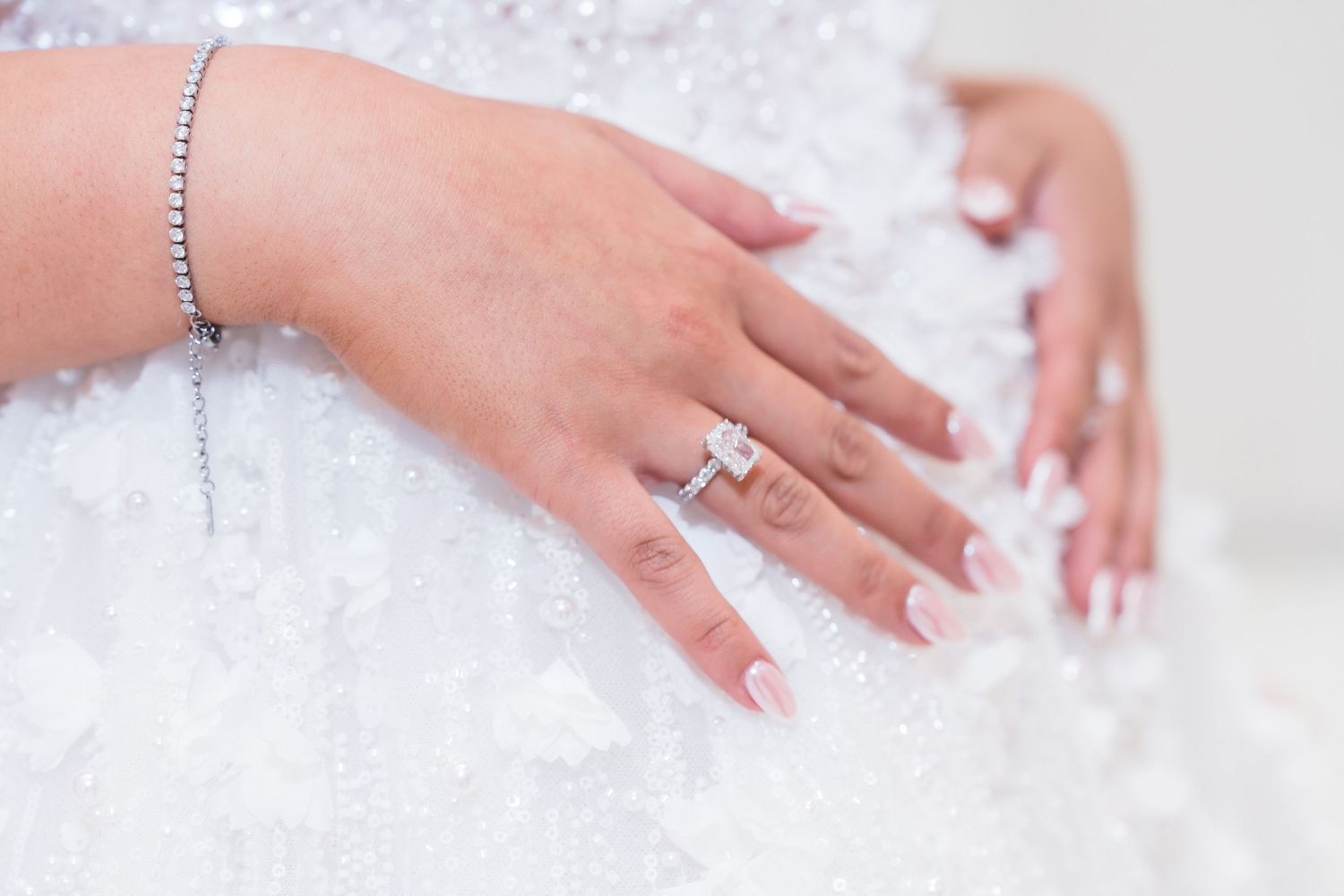 Close-up of bride's hand wearing emerald-cut diamond engagement ring with tennis bracelet on white gown.