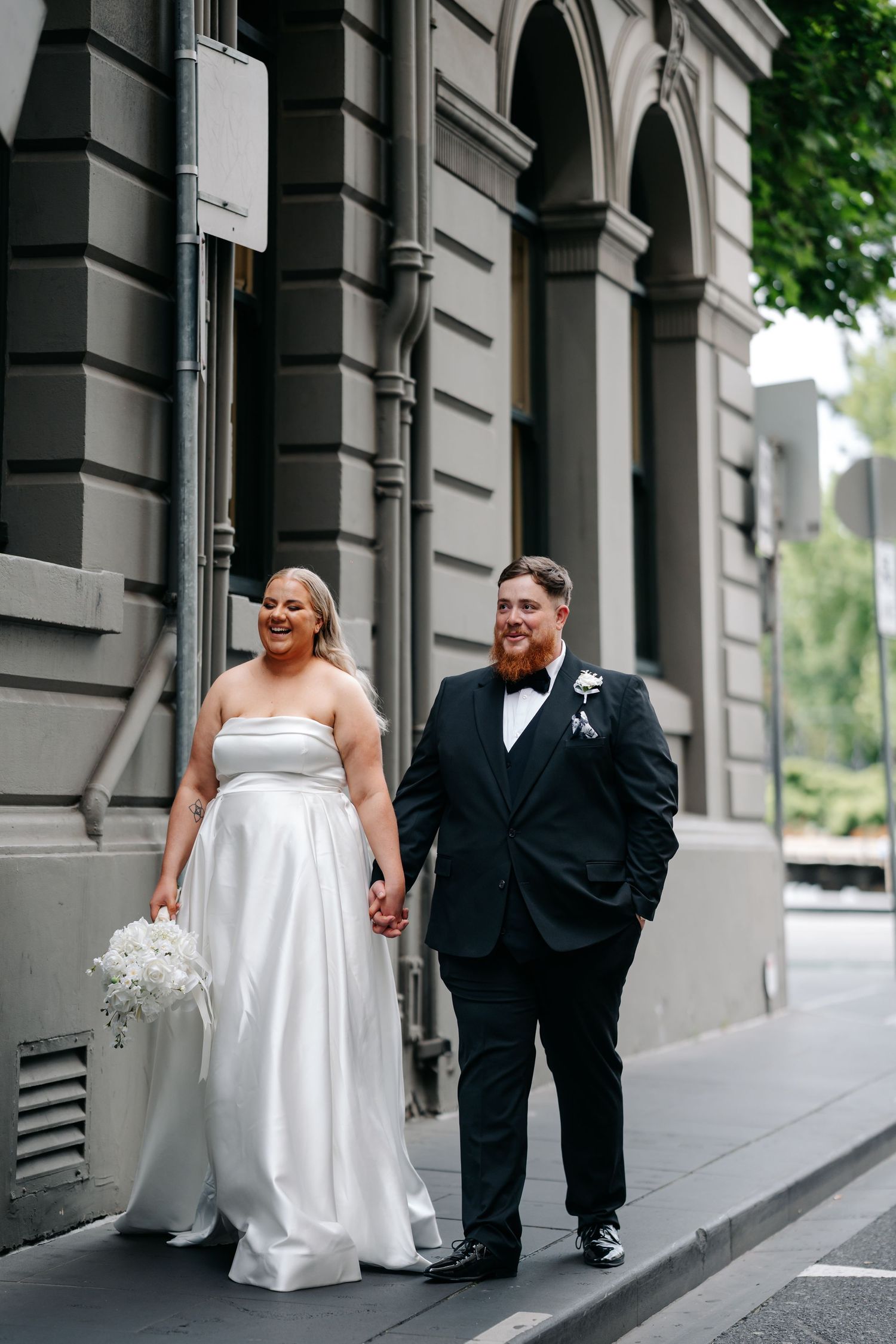 Bride in white strapless gown and groom in black tuxedo walk hand in hand outside a classic stone building.