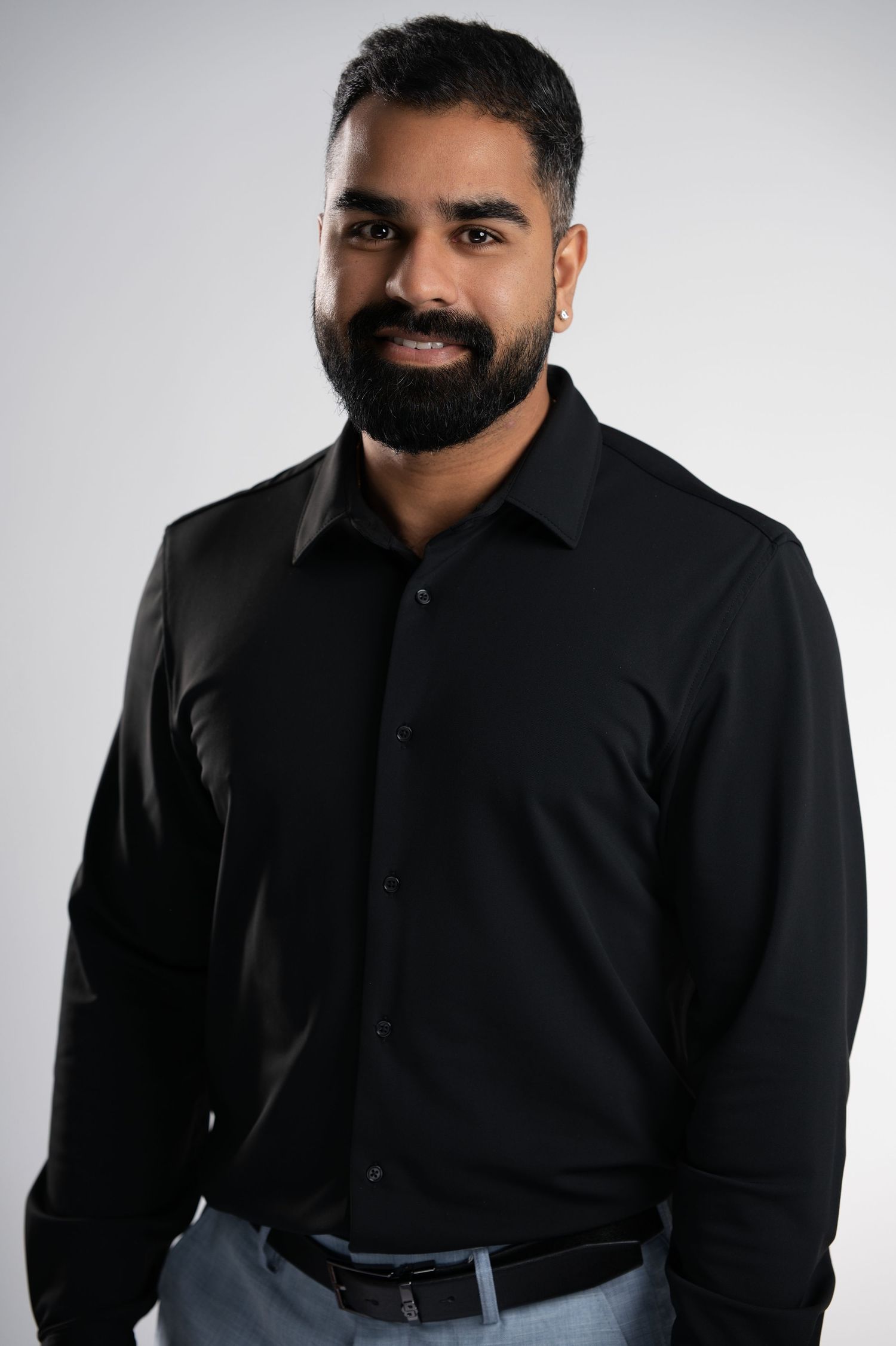 A confident bearded man in a black button-up shirt poses against a light gray studio background.