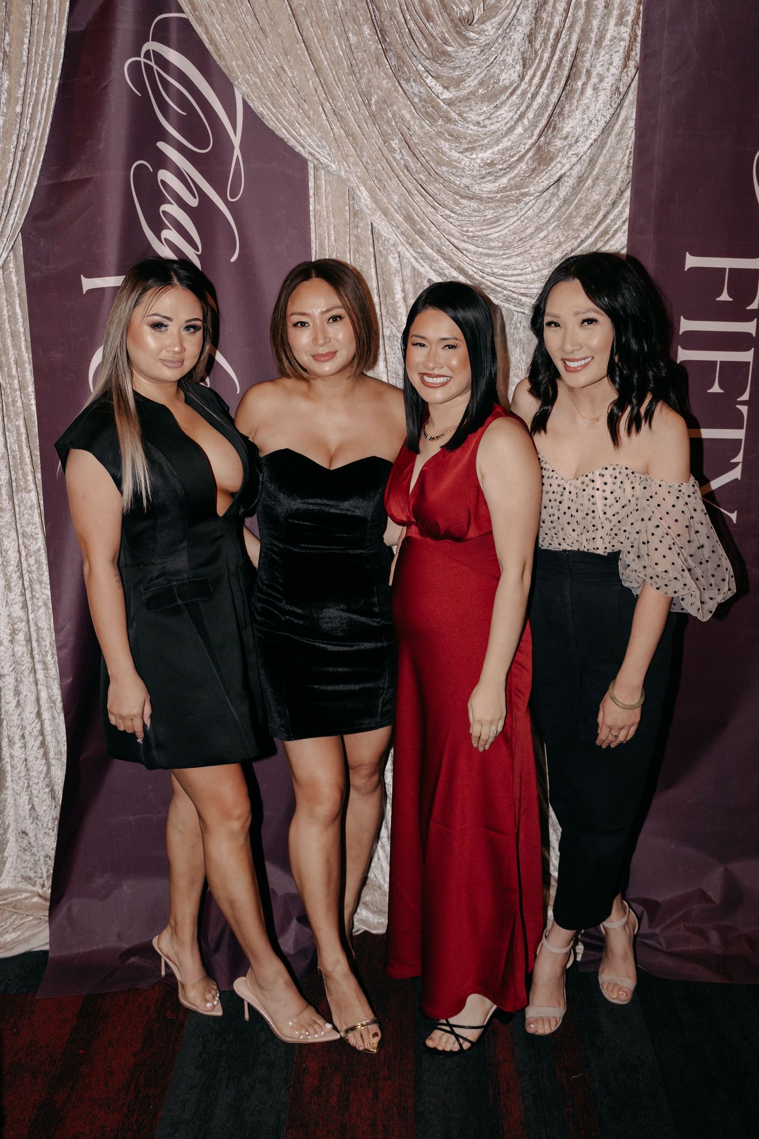 Four women in elegant dresses pose together at a formal event in front of a purple and silver backdrop.