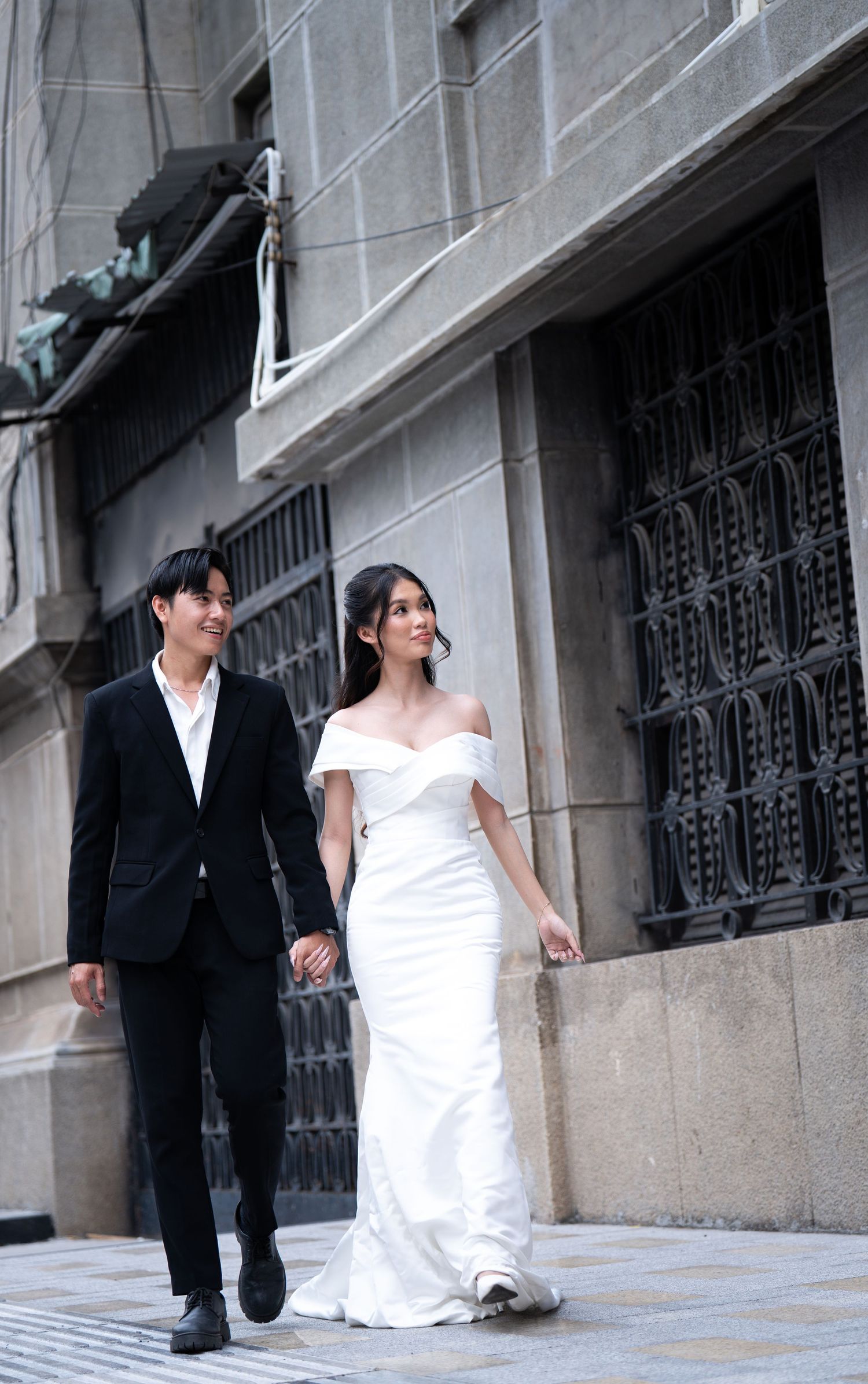A couple in black and white attire walks together along a stone building exterior.