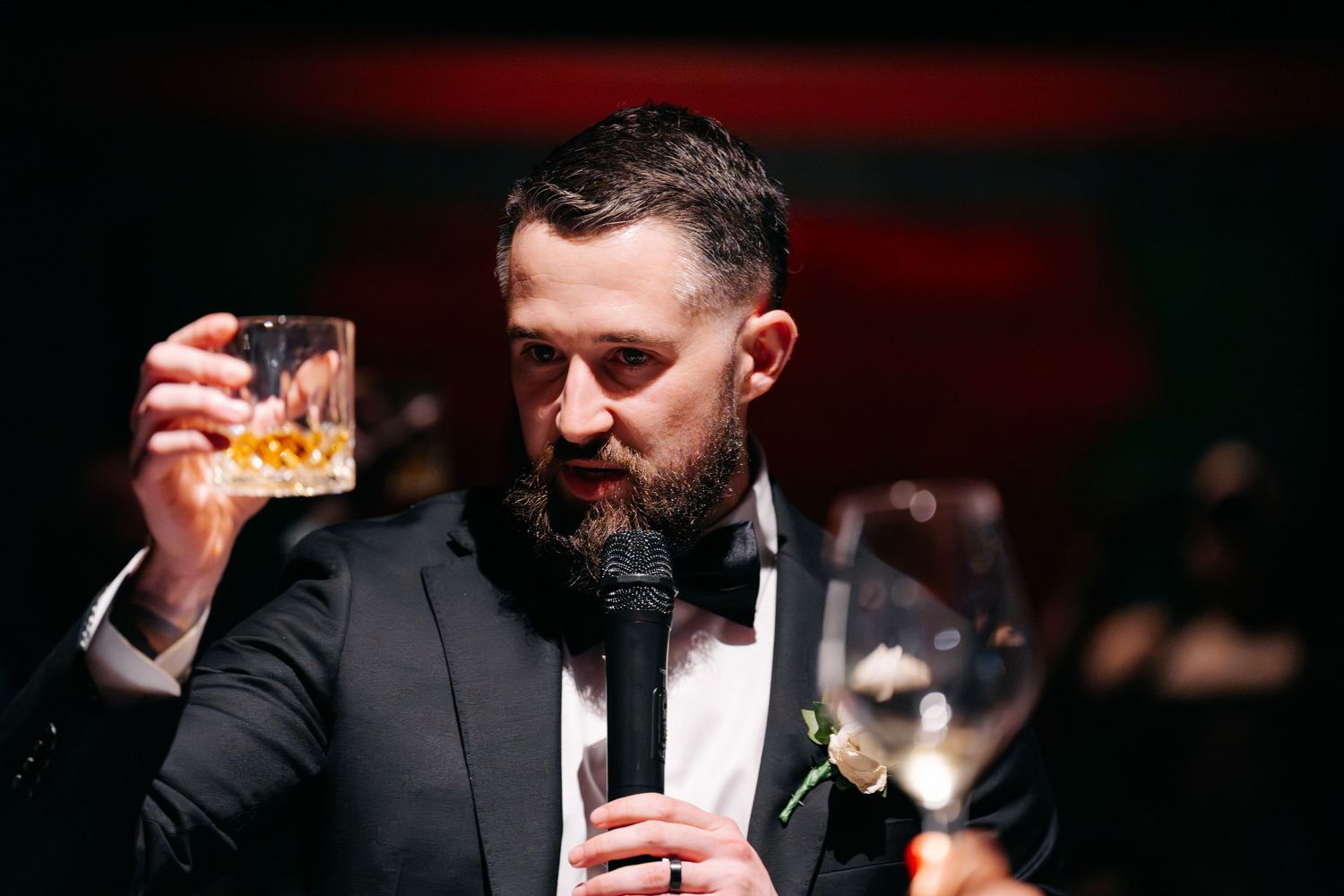 A man in a tuxedo raises a cocktail glass while speaking into a microphone at an evening event.