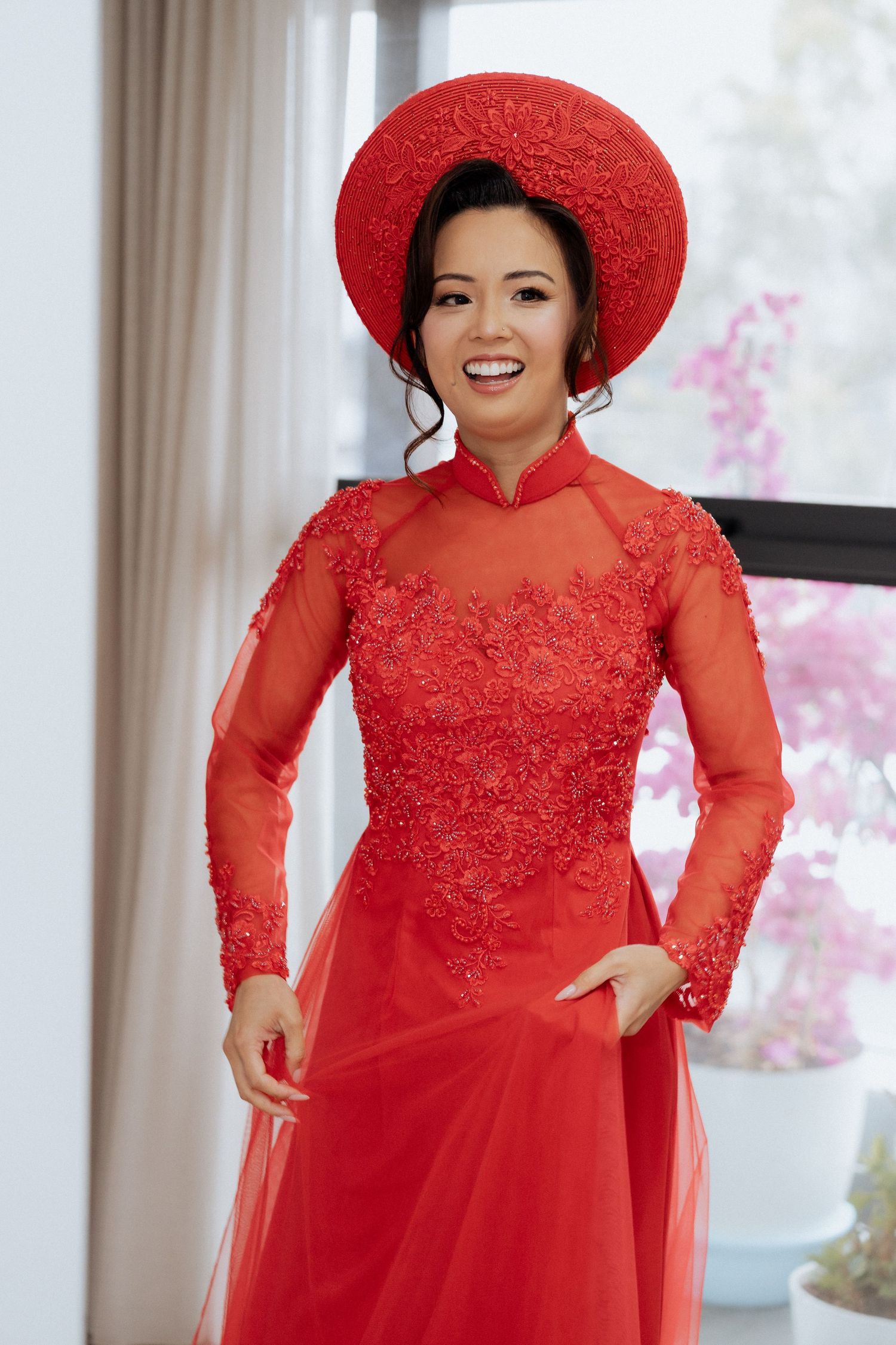 Smiling woman in red Vietnamese Ao Dai dress with lace bodice and matching red non la hat, standing indoors.