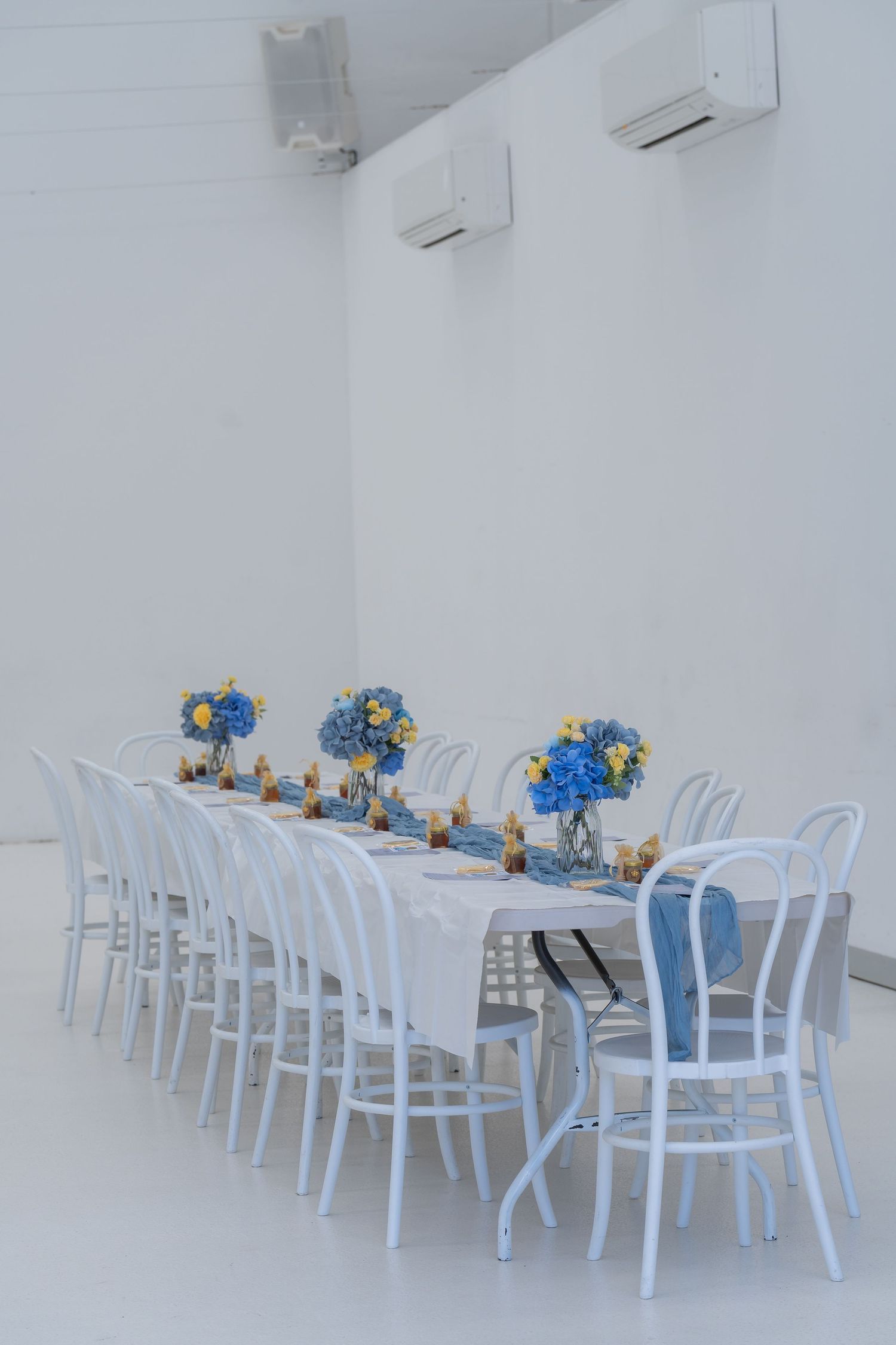Elegant white banquet table with blue and yellow floral centerpieces, surrounded by white chairs in a bright white room.