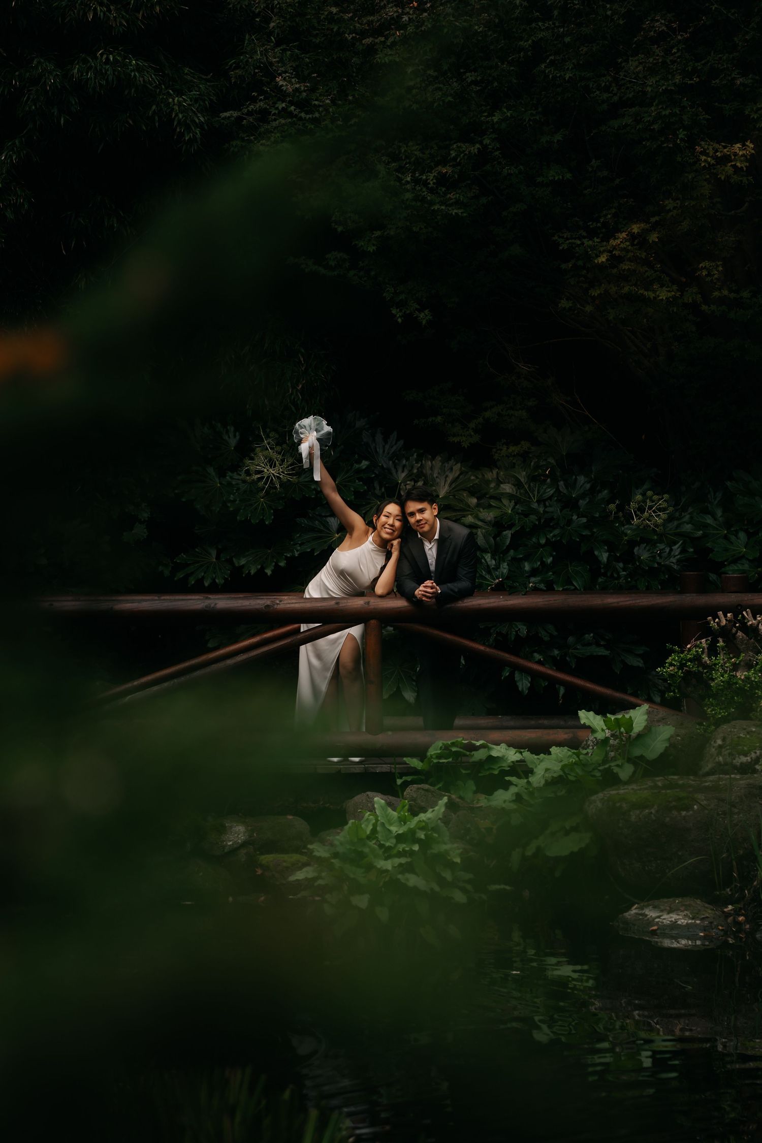 Joyful bride raising bouquet on a wooden bridge surrounded by lush green forest with groom.