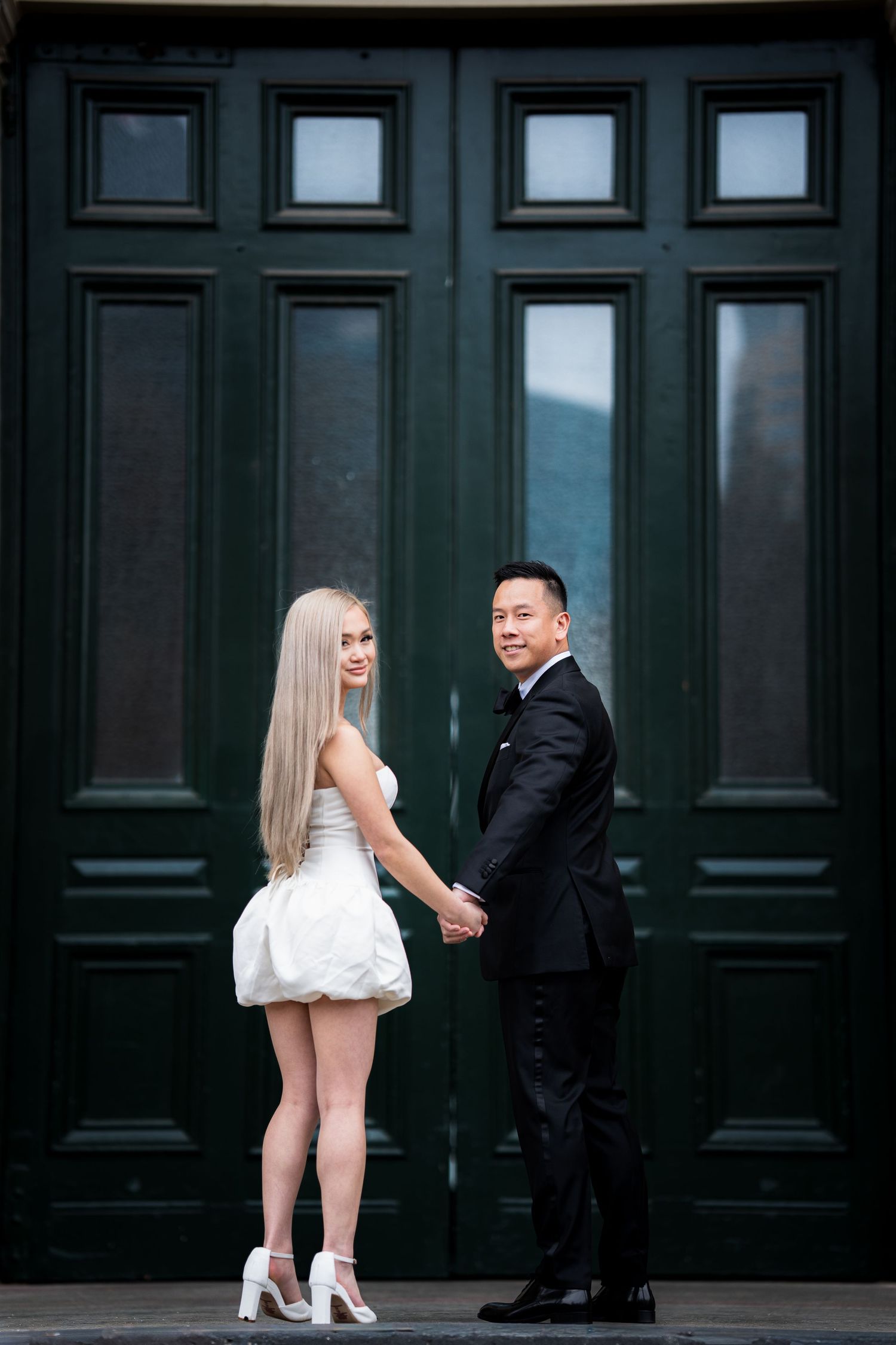 A couple poses in formal wear against a large dark green paneled door.