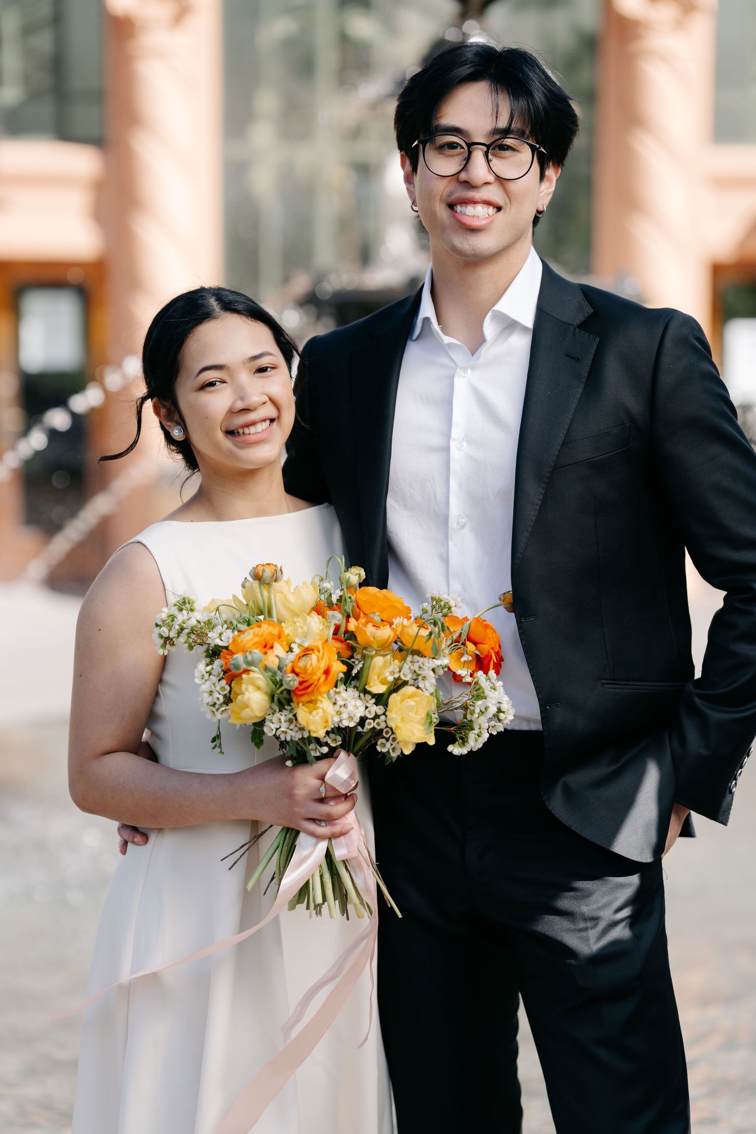 A couple in formal attire poses with a vibrant orange and yellow bouquet.
