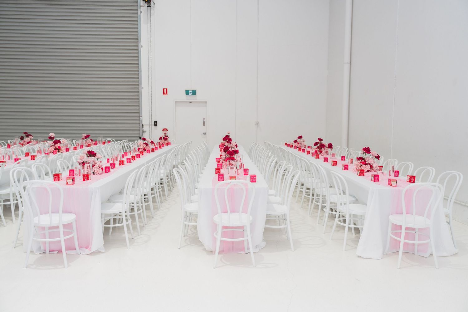 Elegant event venue with long white tables, pink and red floral centerpieces, and white chairs in a bright white room.