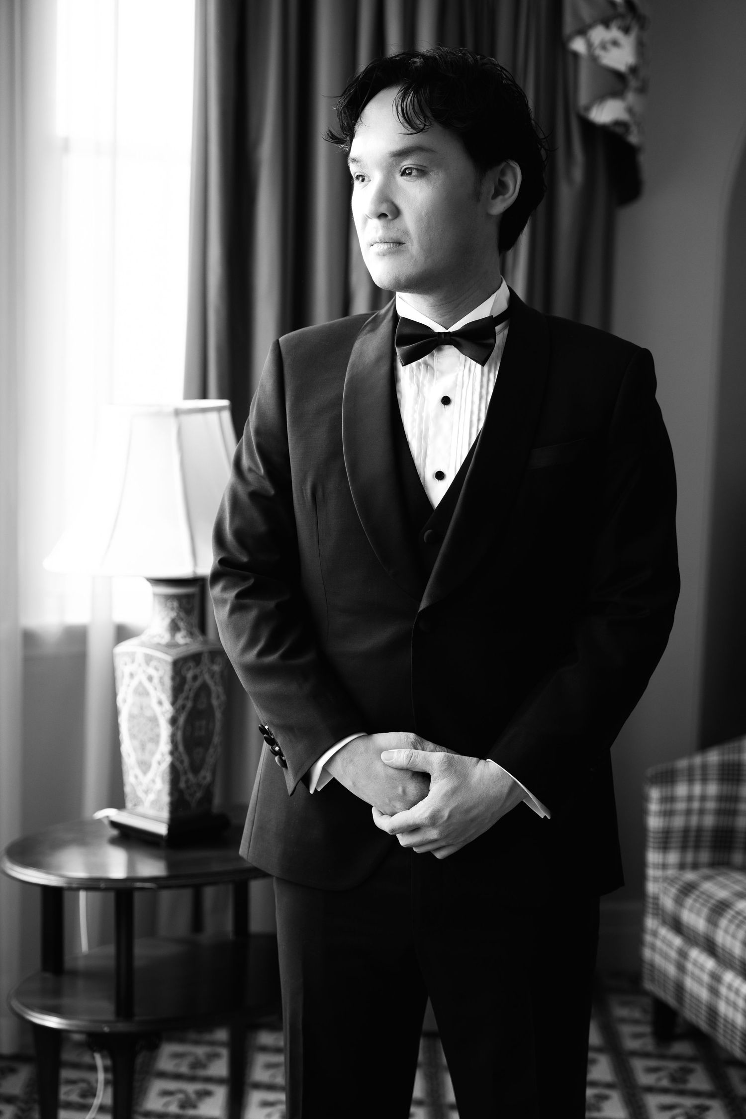 Black and white portrait of a young man in a tuxedo with bow tie standing by a window in an elegant room.