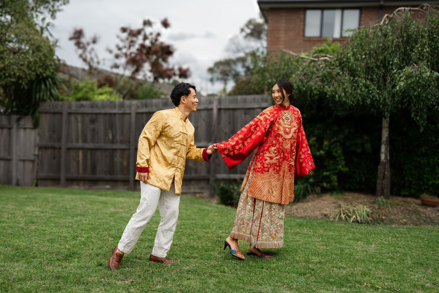 Couple in traditional Asian attire holding hands and dancing joyfully in a lush green backyard garden.