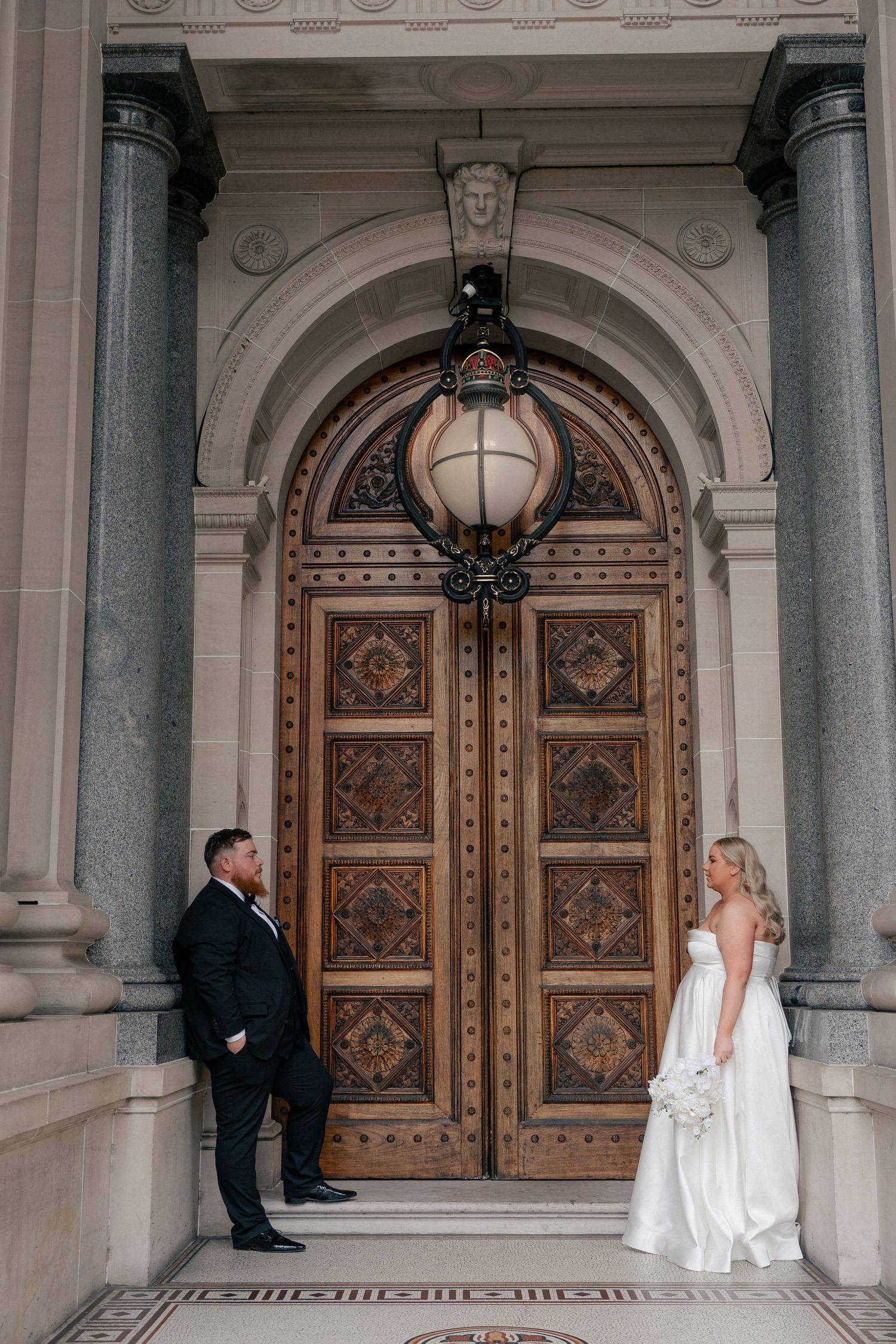 Bride and groom pose apart before ornate wooden doors of a grand stone building on their wedding day.