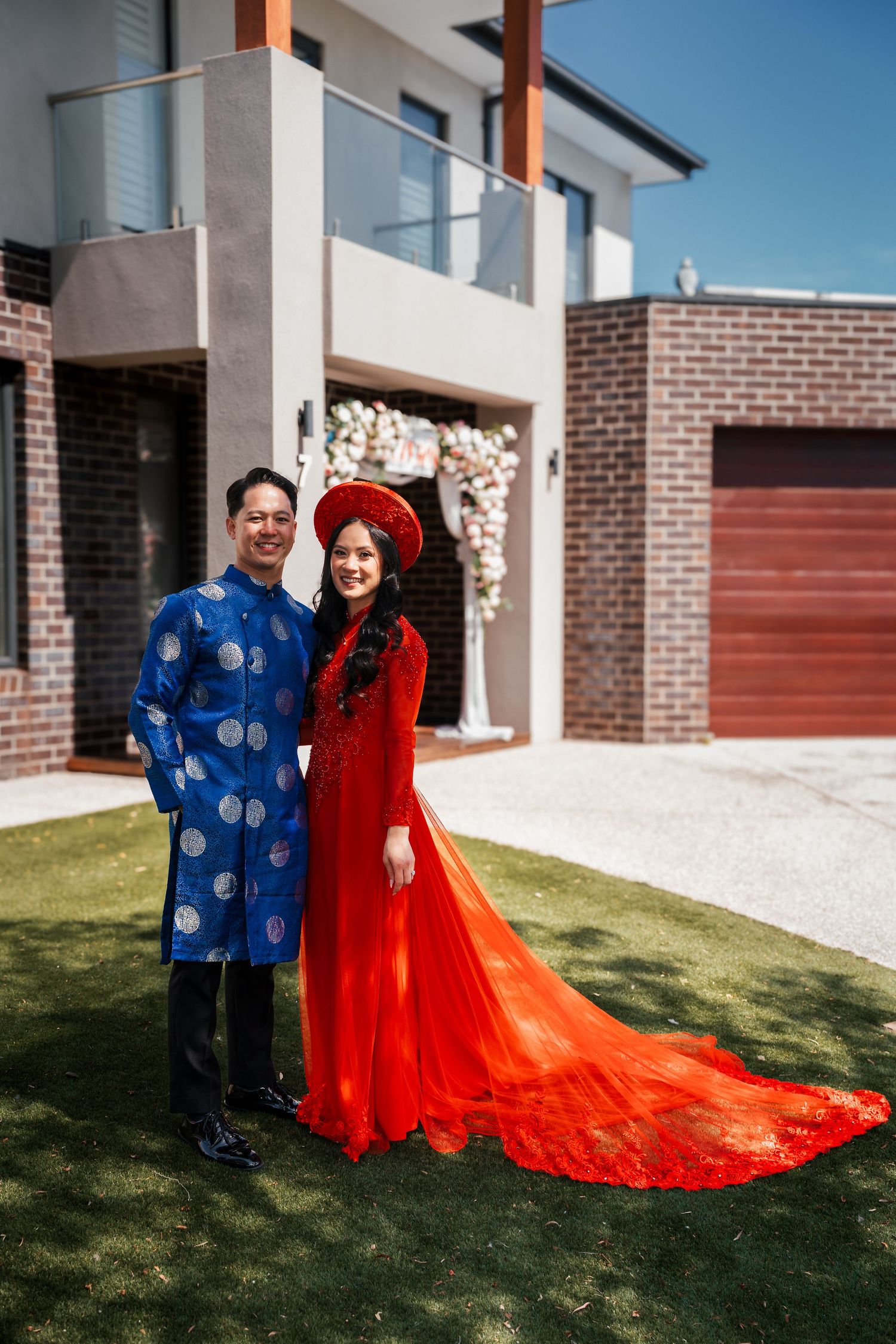Vietnamese wedding couple in traditional ao dai standing in front of modern brick home.