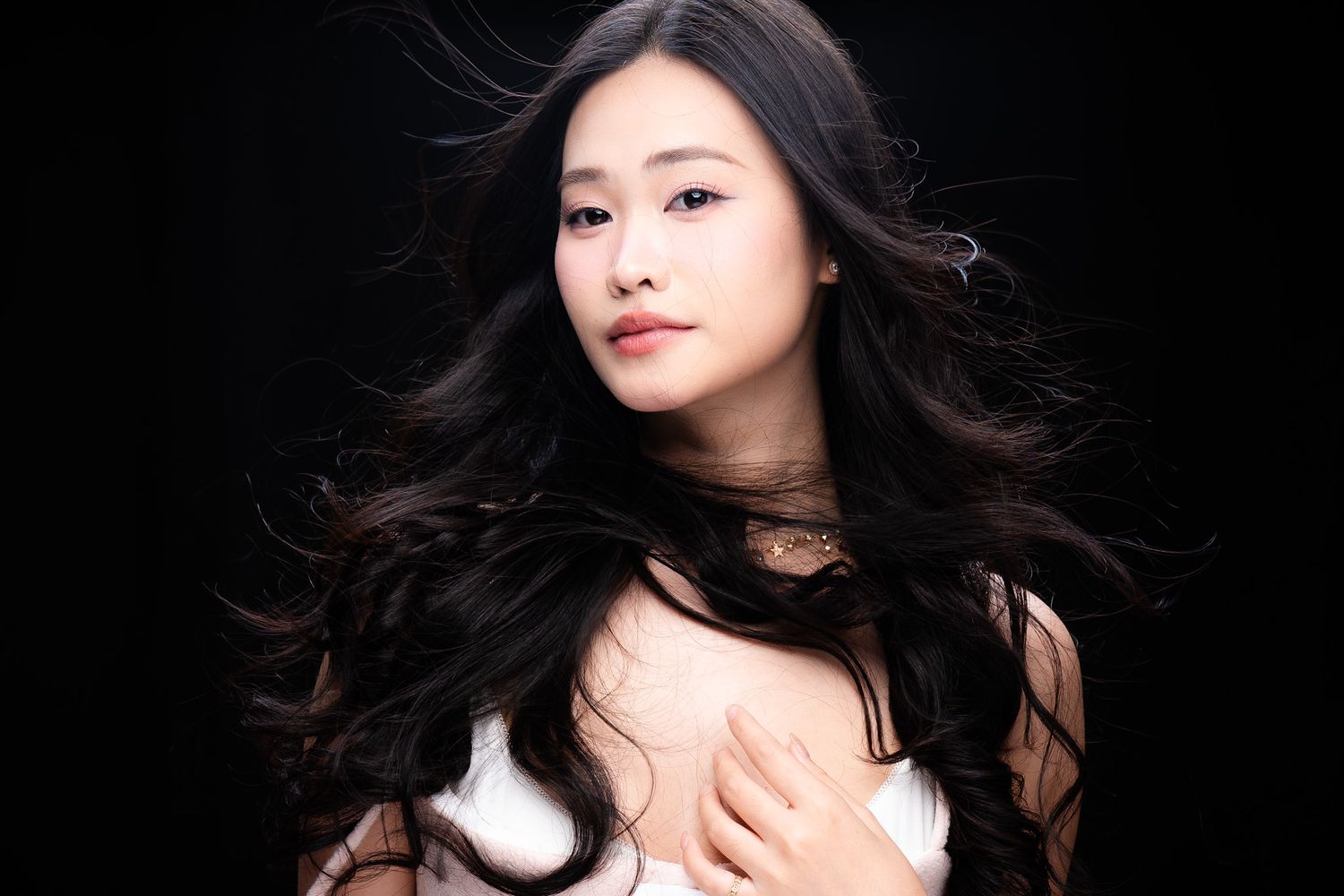 Young Asian woman with long wavy black hair posing elegantly against a dark background in a white outfit.