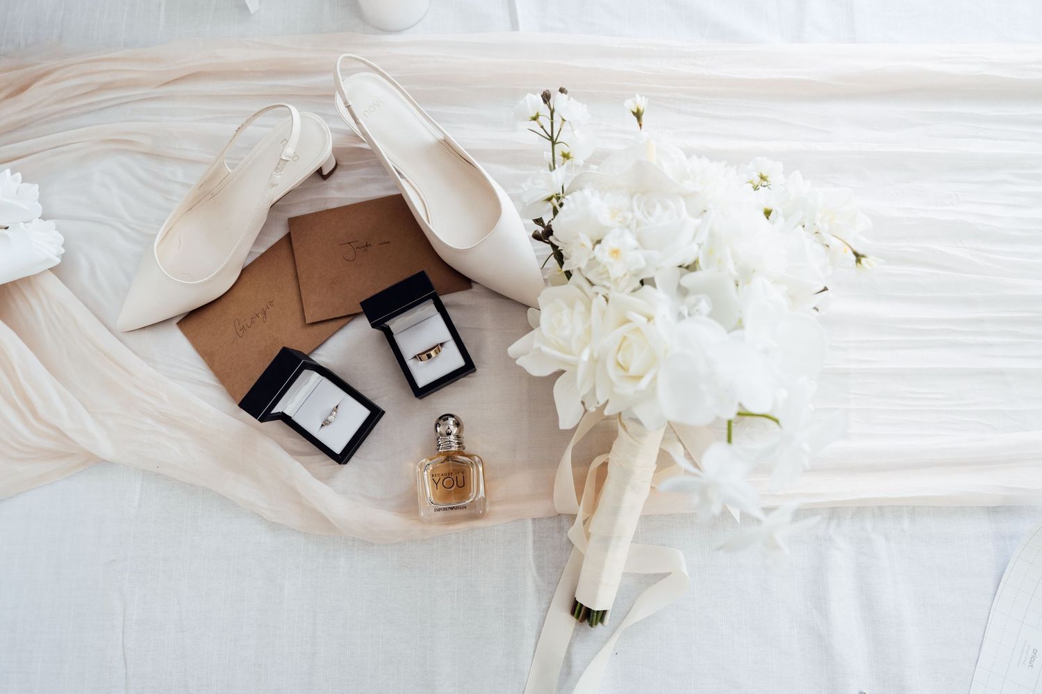 Elegant wedding flat lay featuring bridal shoes, ring boxes, perfume bottle, and white floral bouquet on soft fabric.
