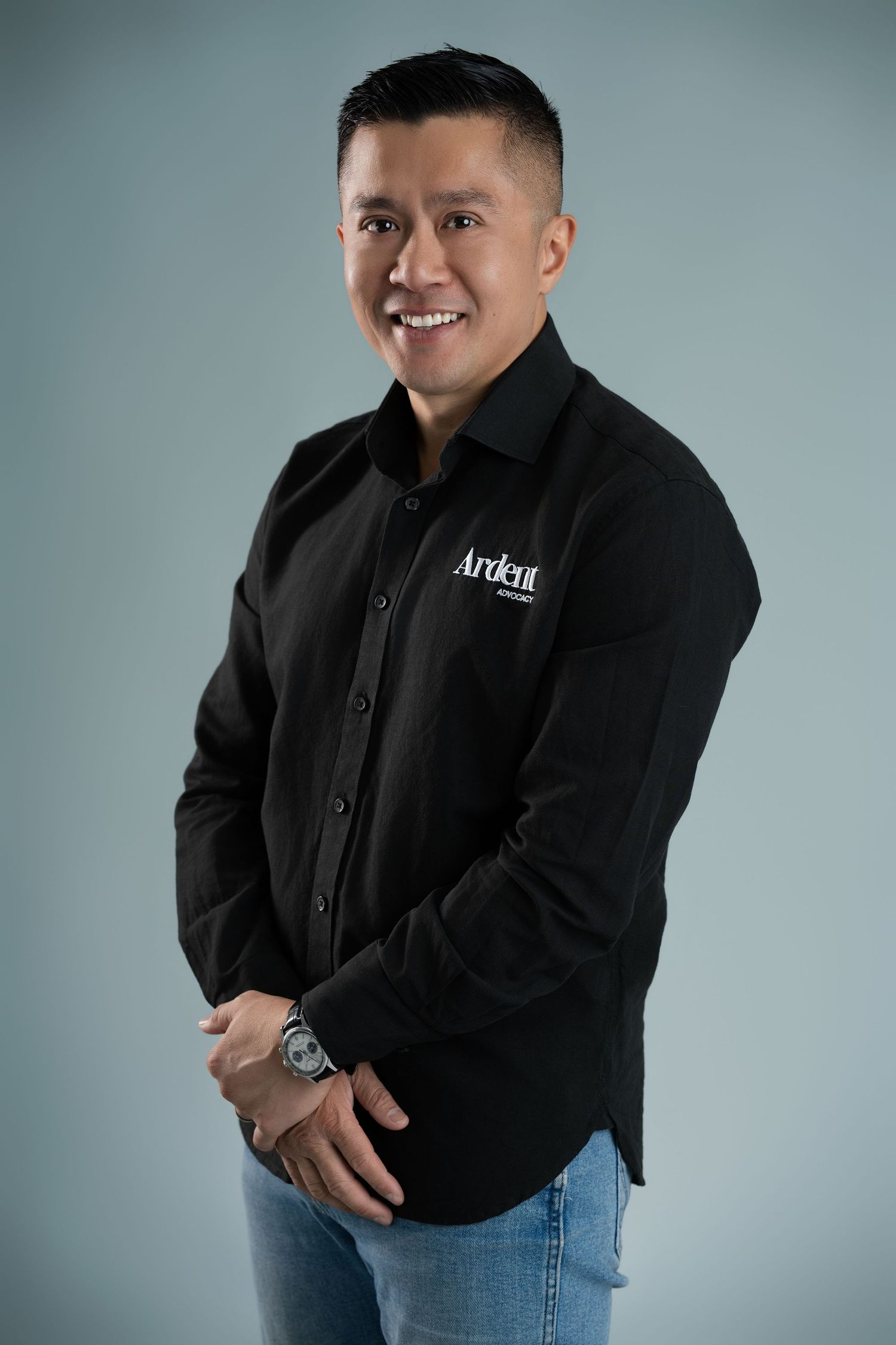 Man in black Ardent logo shirt smiling, wearing a watch, posing confidently against a gray background.