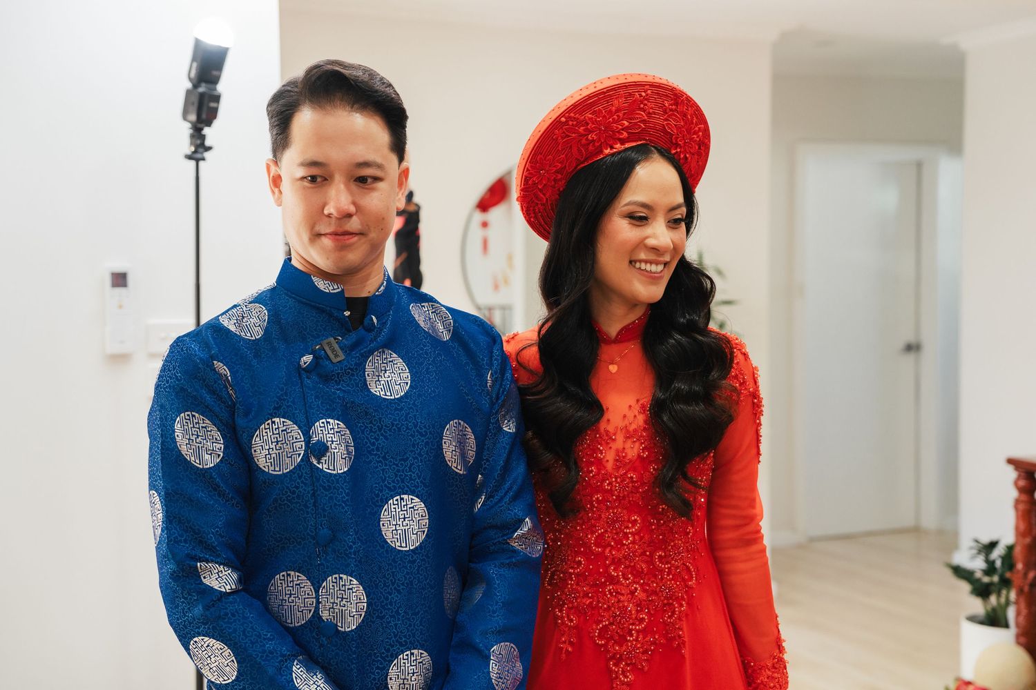 A Vietnamese couple in traditional ao dai attire, man in blue and woman in red, smiling during a wedding ceremony.