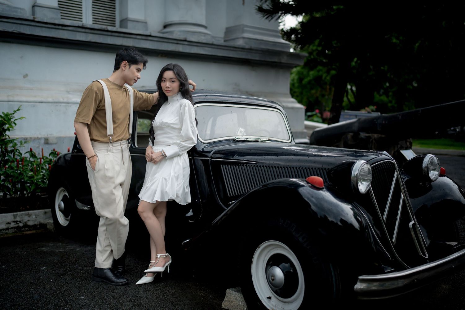 Elegant couple in vintage attire posing beside a classic black retro car in an outdoor setting.