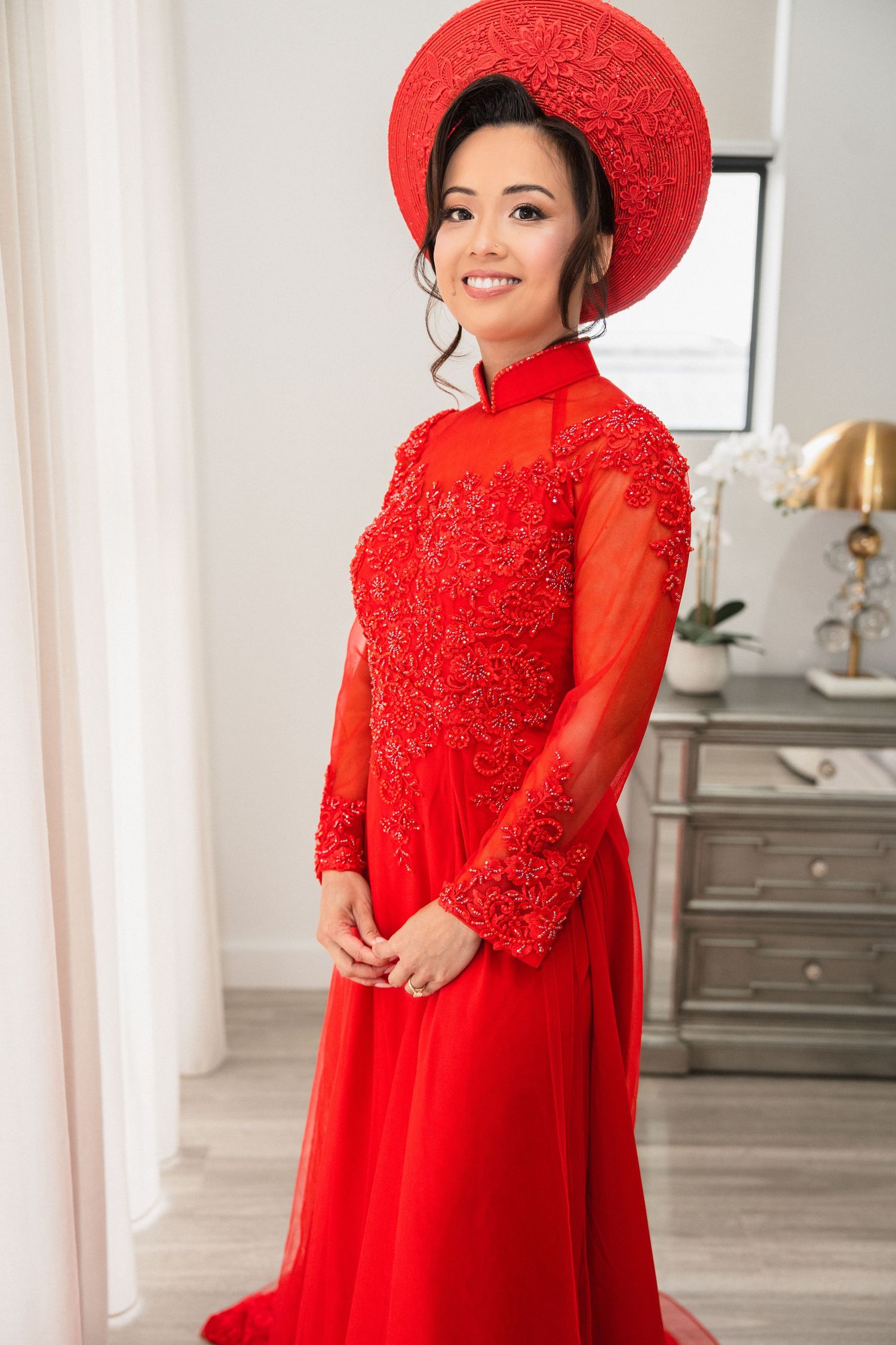 Smiling woman wearing a traditional red Vietnamese Ao Dai gown with lace detailing and matching wide-brim Non La hat.