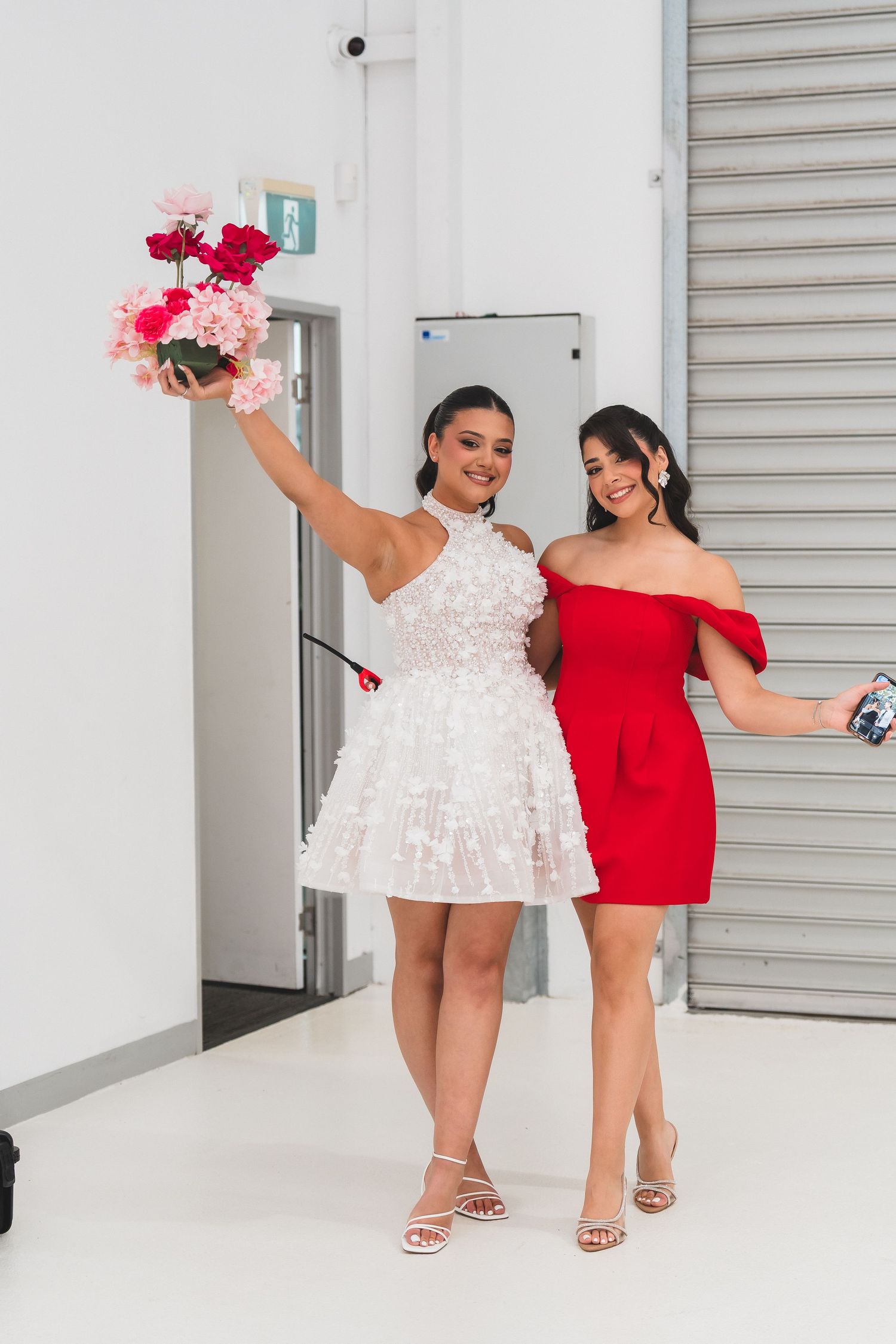 Two young women in white and red dresses posing with pink and red flowers at a formal event.