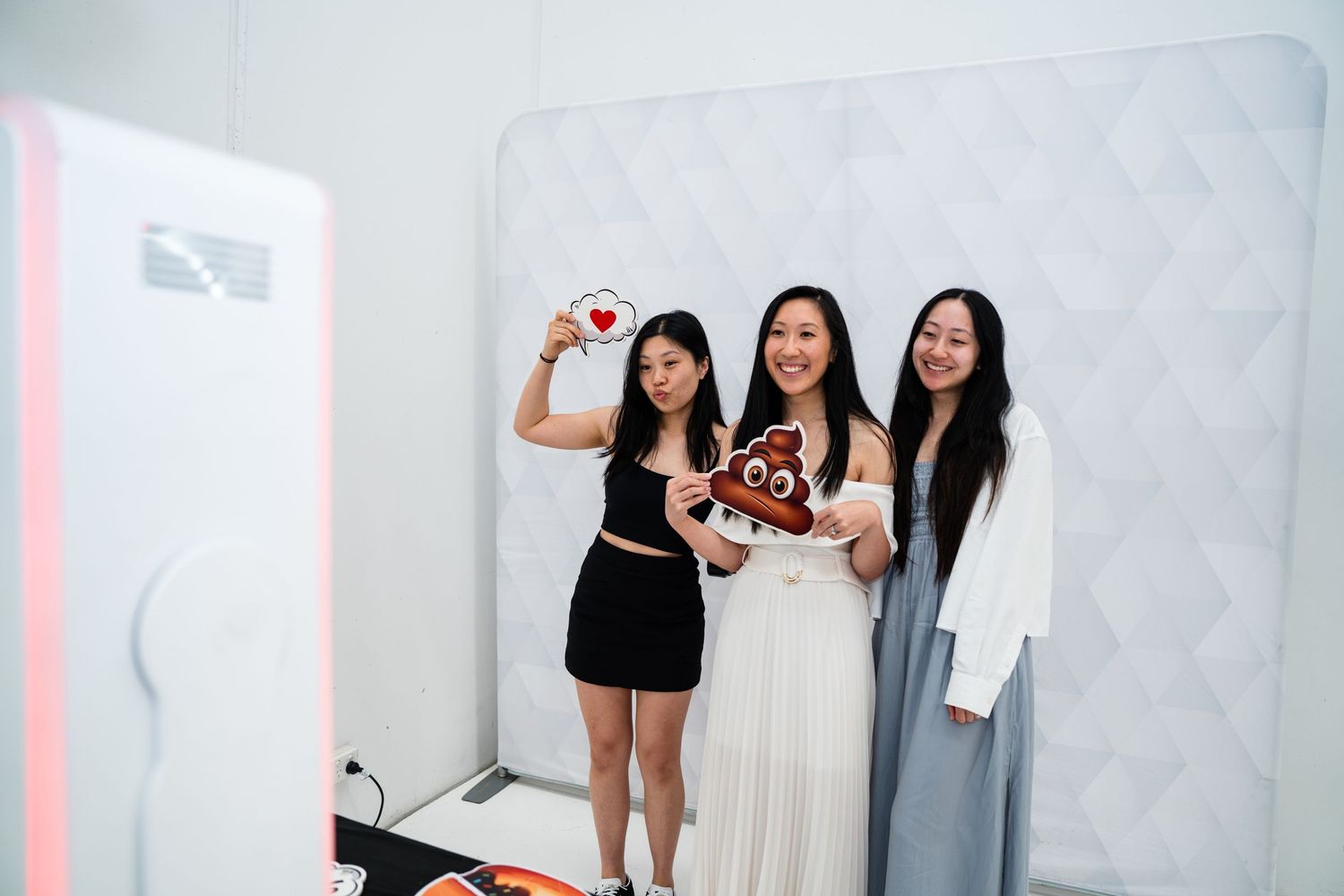 Three women smile and pose with emoji pillows in front of a photo booth at an indoor event.