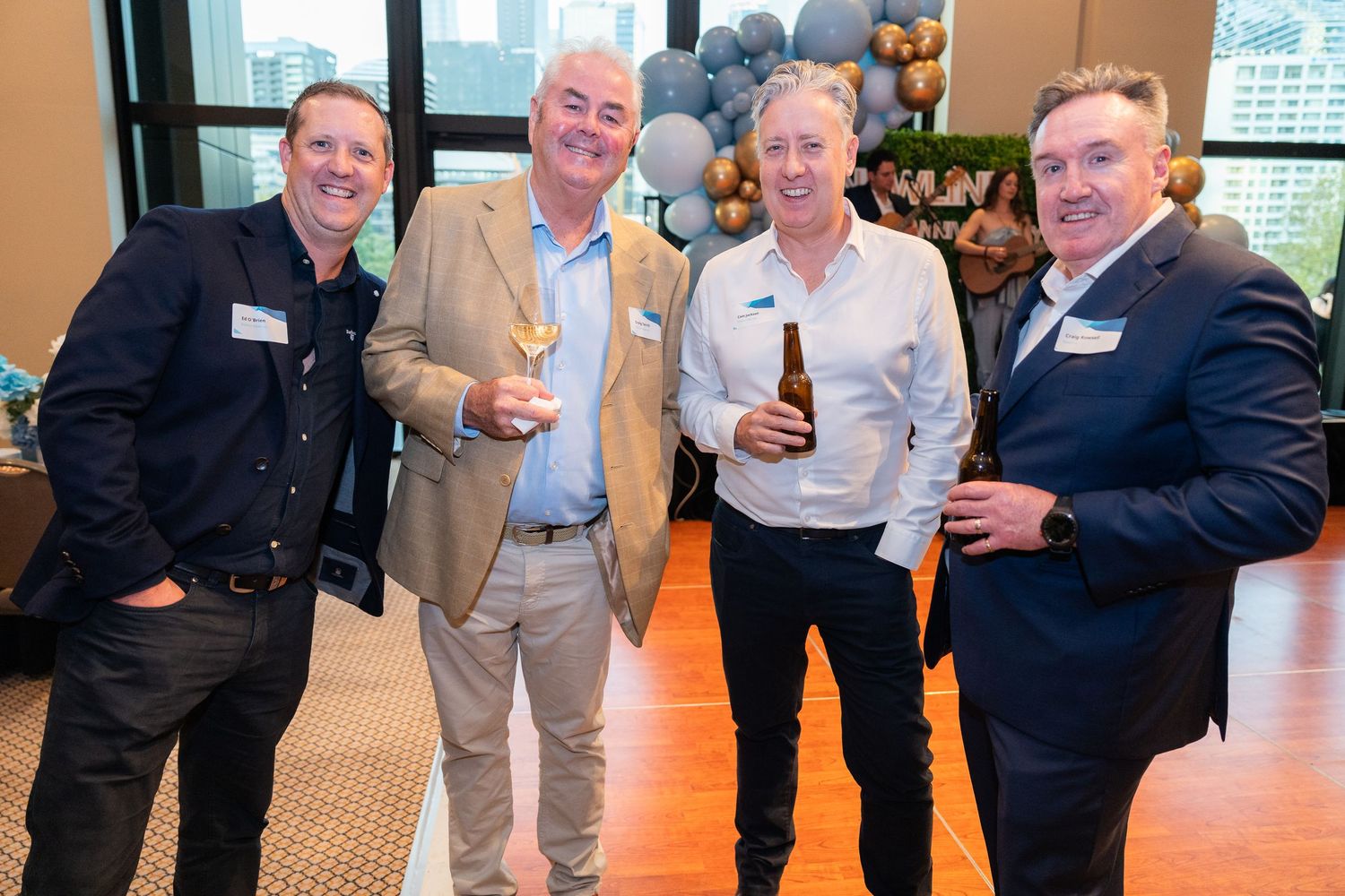 Four smiling men posing together at a corporate networking event, holding drinks with balloon decorations behind them.