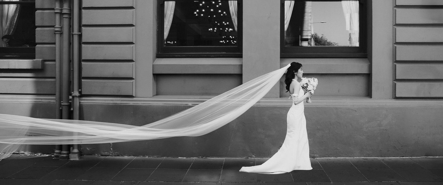 Bride in white gown with long flowing veil on city sidewalk, black and white wedding photography.