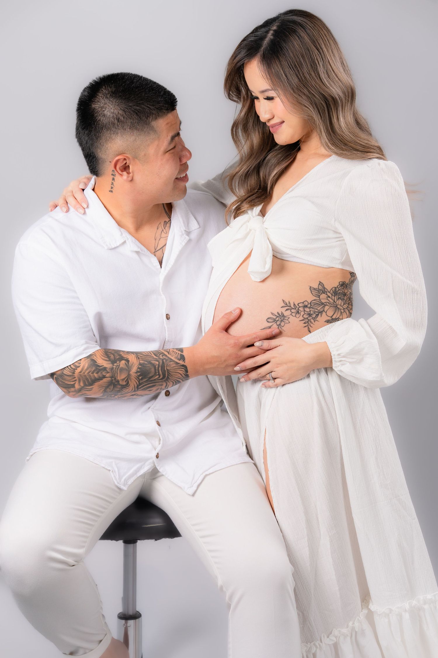 Tattooed couple in white outfits posing for maternity photo, man seated on stool touching pregnant woman's belly.