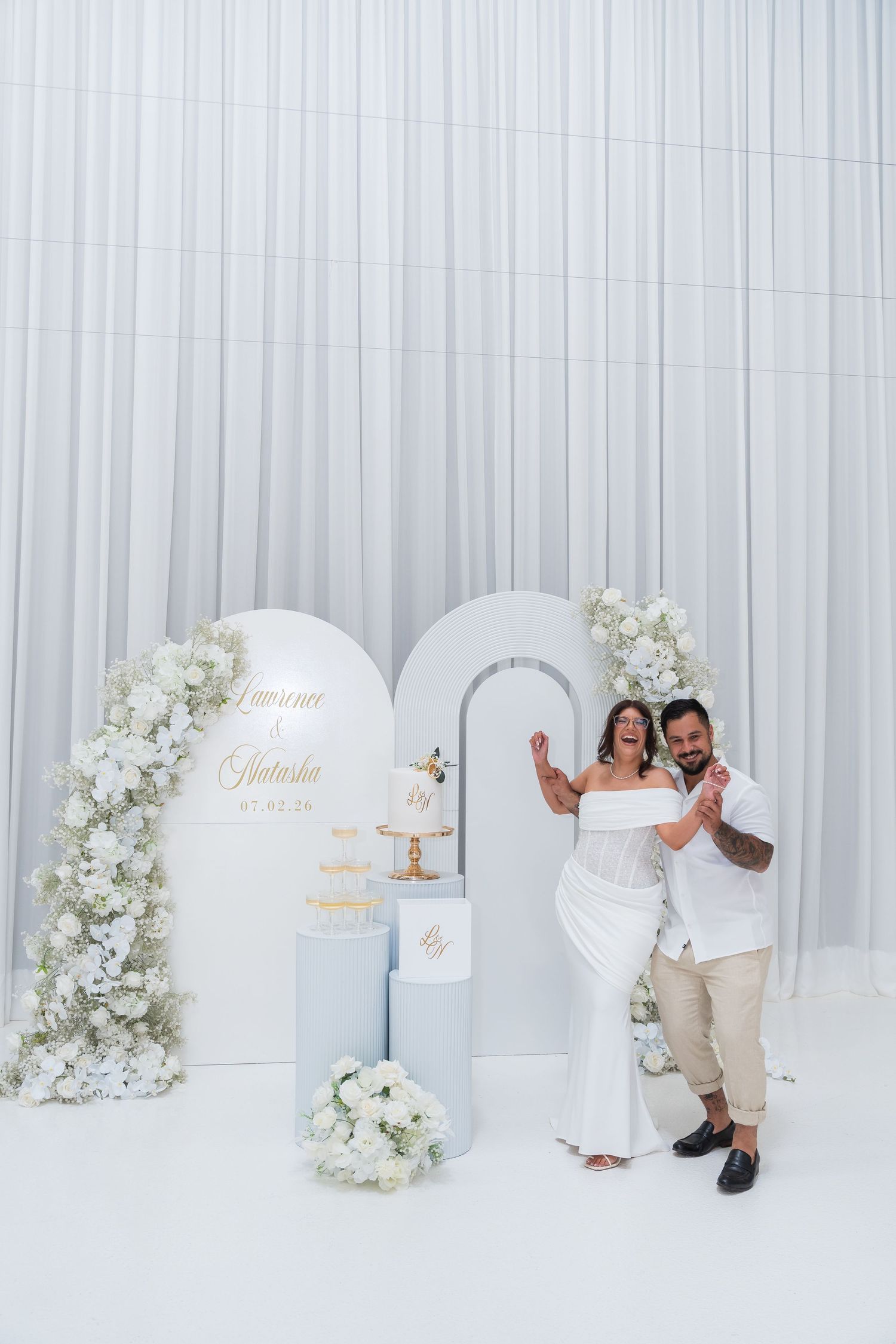 Couple celebrating wedding in elegant white venue with floral arch, tiered cake display, and white drape backdrop.