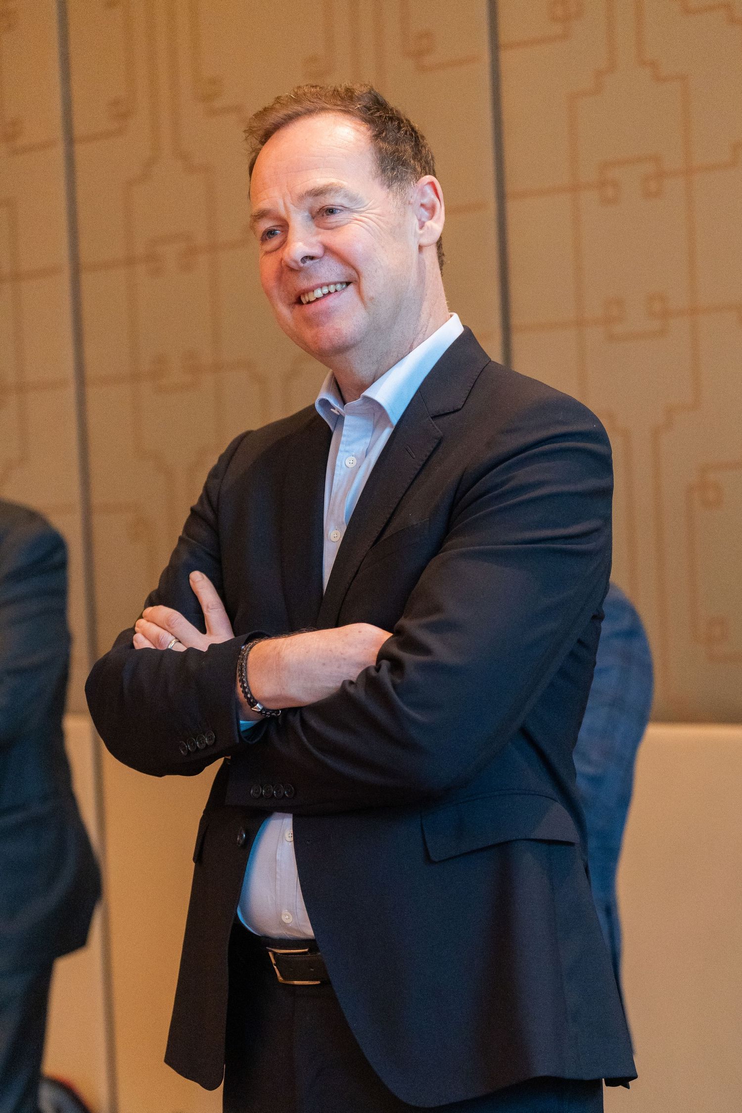 Smiling businessman in dark suit with arms crossed standing in modern conference room.