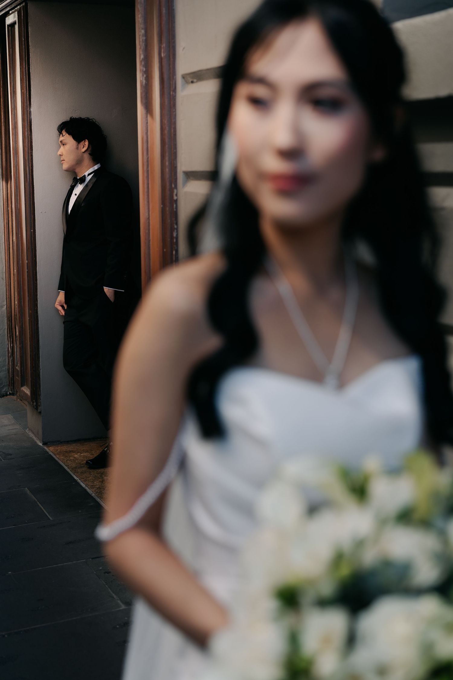 Bride in white gown holds bouquet in foreground as groom in black tuxedo leans against wall in background.