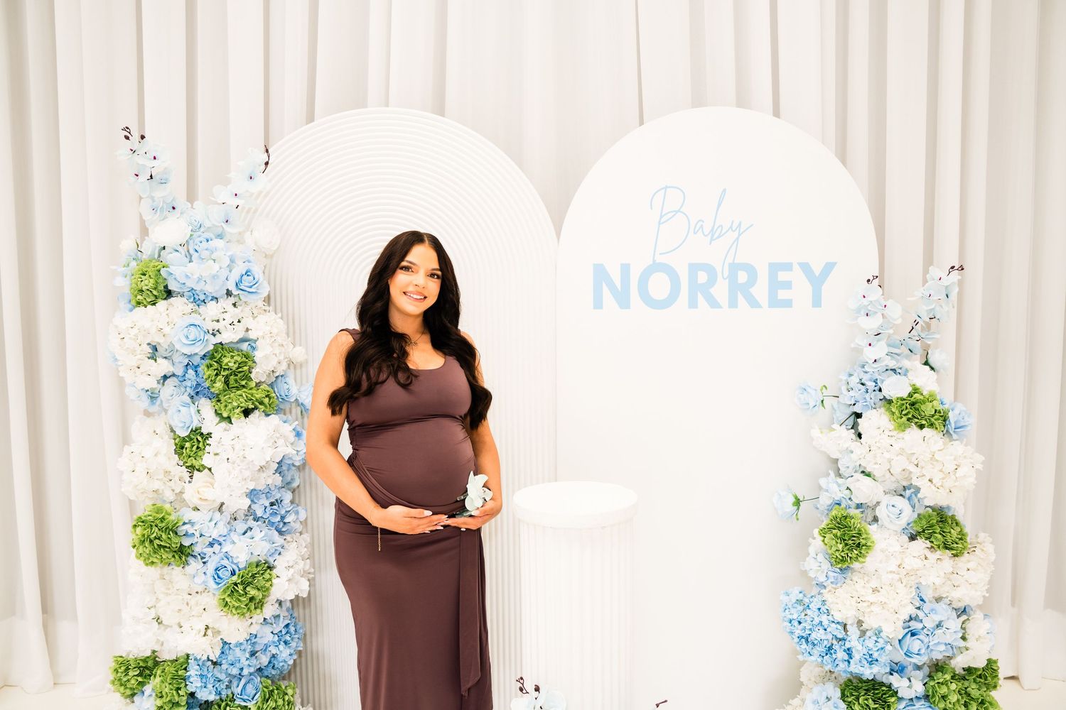 Pregnant woman in brown dress poses at Baby Norrey baby shower with blue and white floral arrangements.