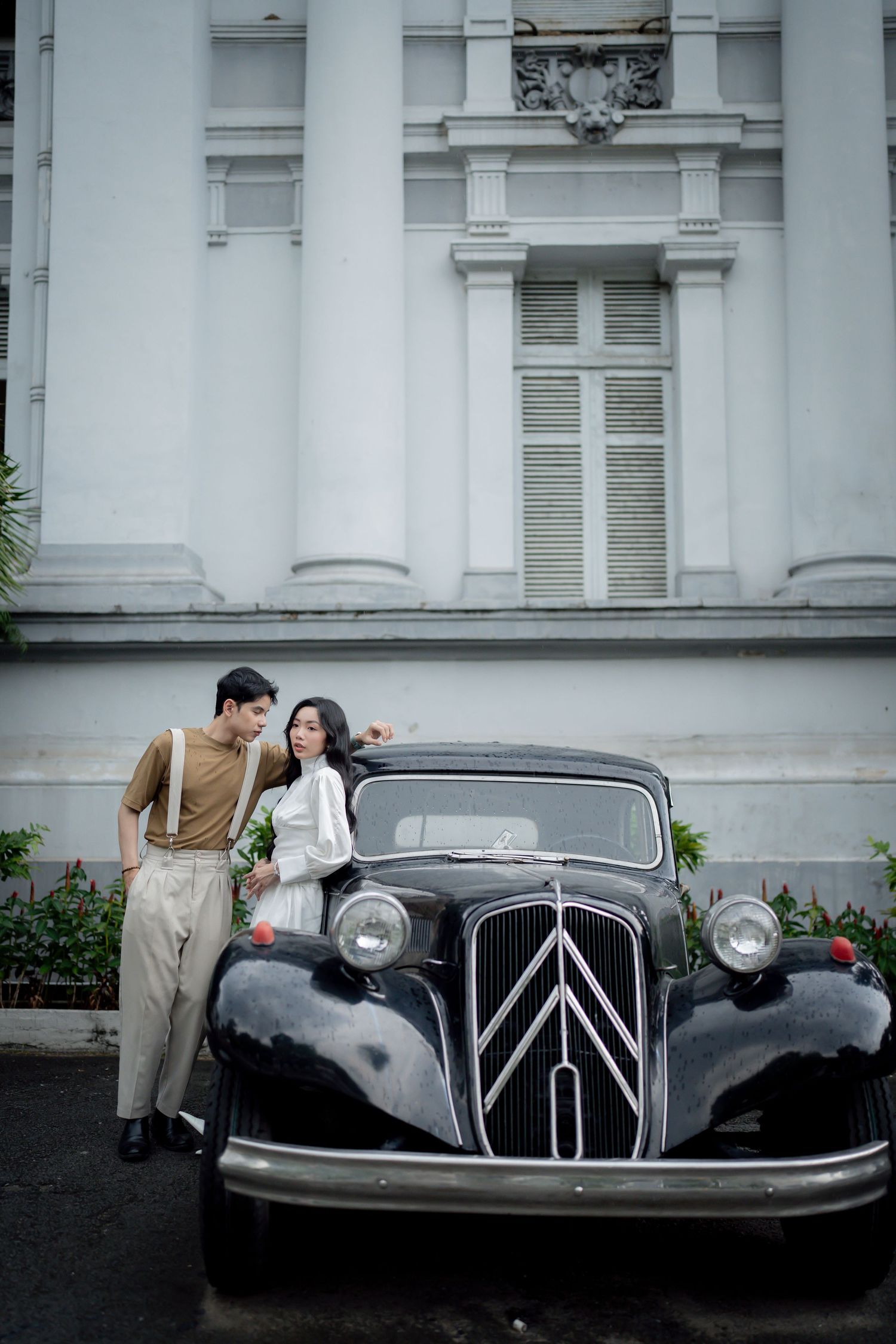 A vintage black car is parked in front of a white colonial building during a romantic photoshoot.