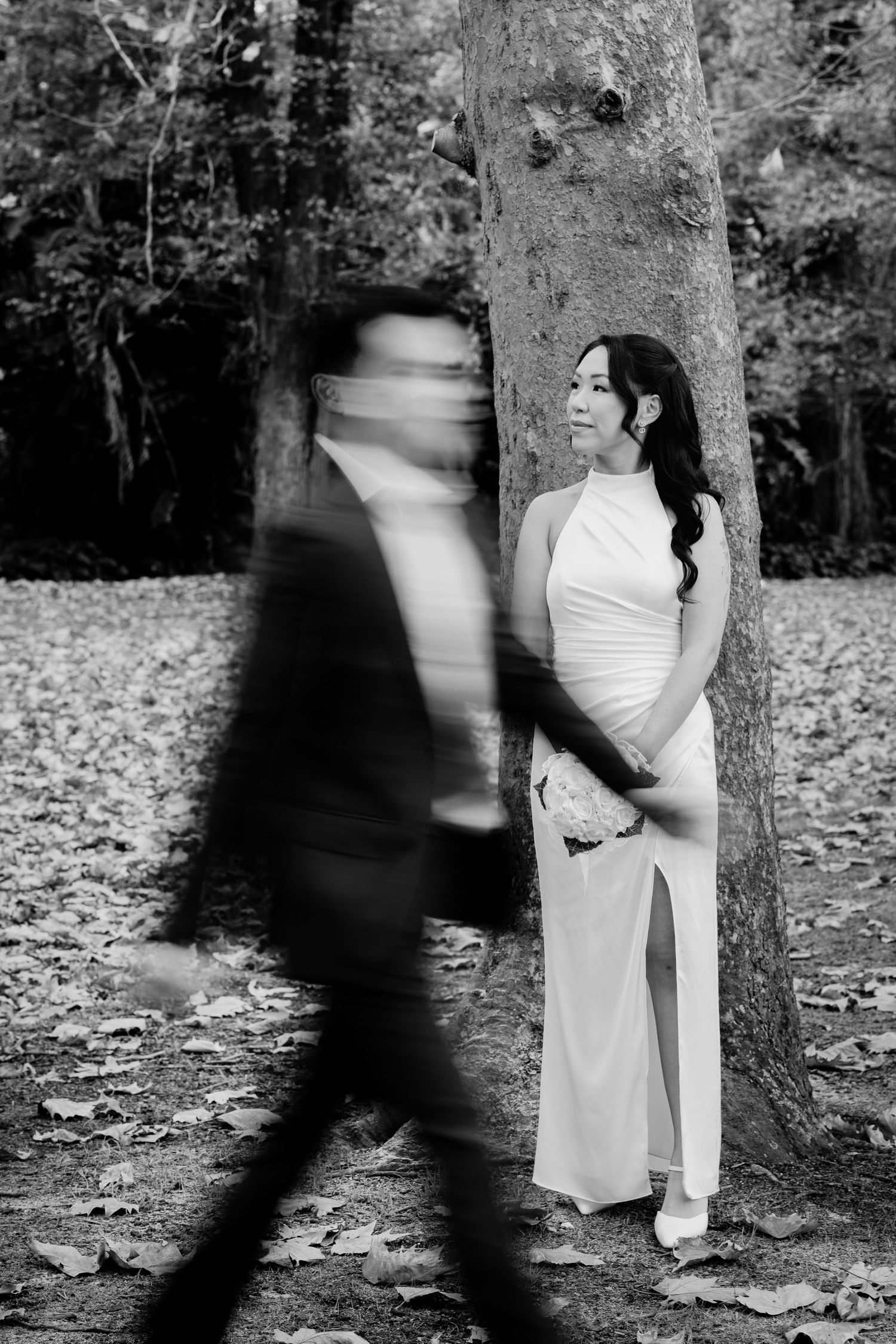Black and white wedding photo of bride leaning against tree while groom walks past with motion blur in autumn forest.